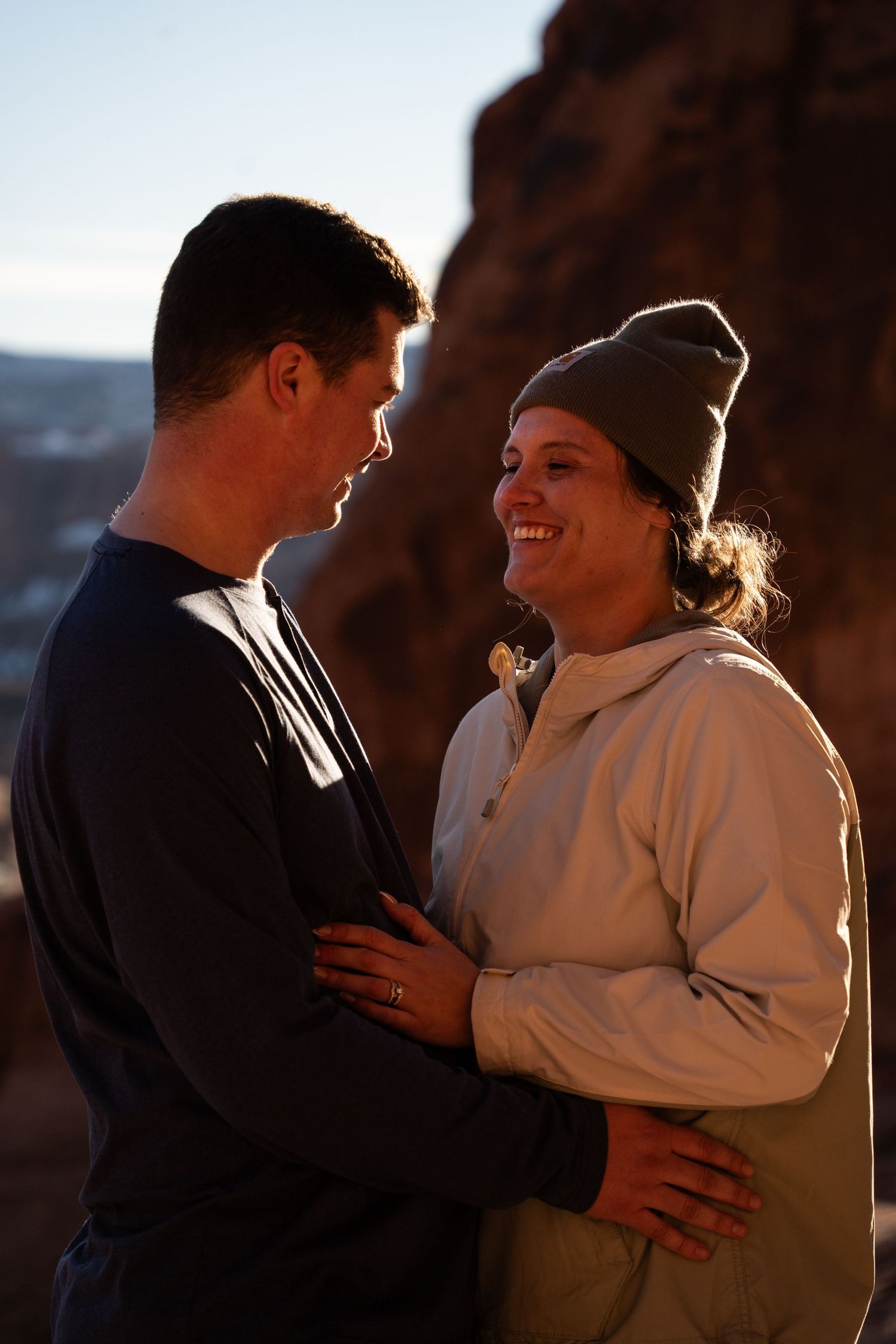 Couple embracing and smiling at sunset near red rock formations, woman wearing gray beanie and hoodie.