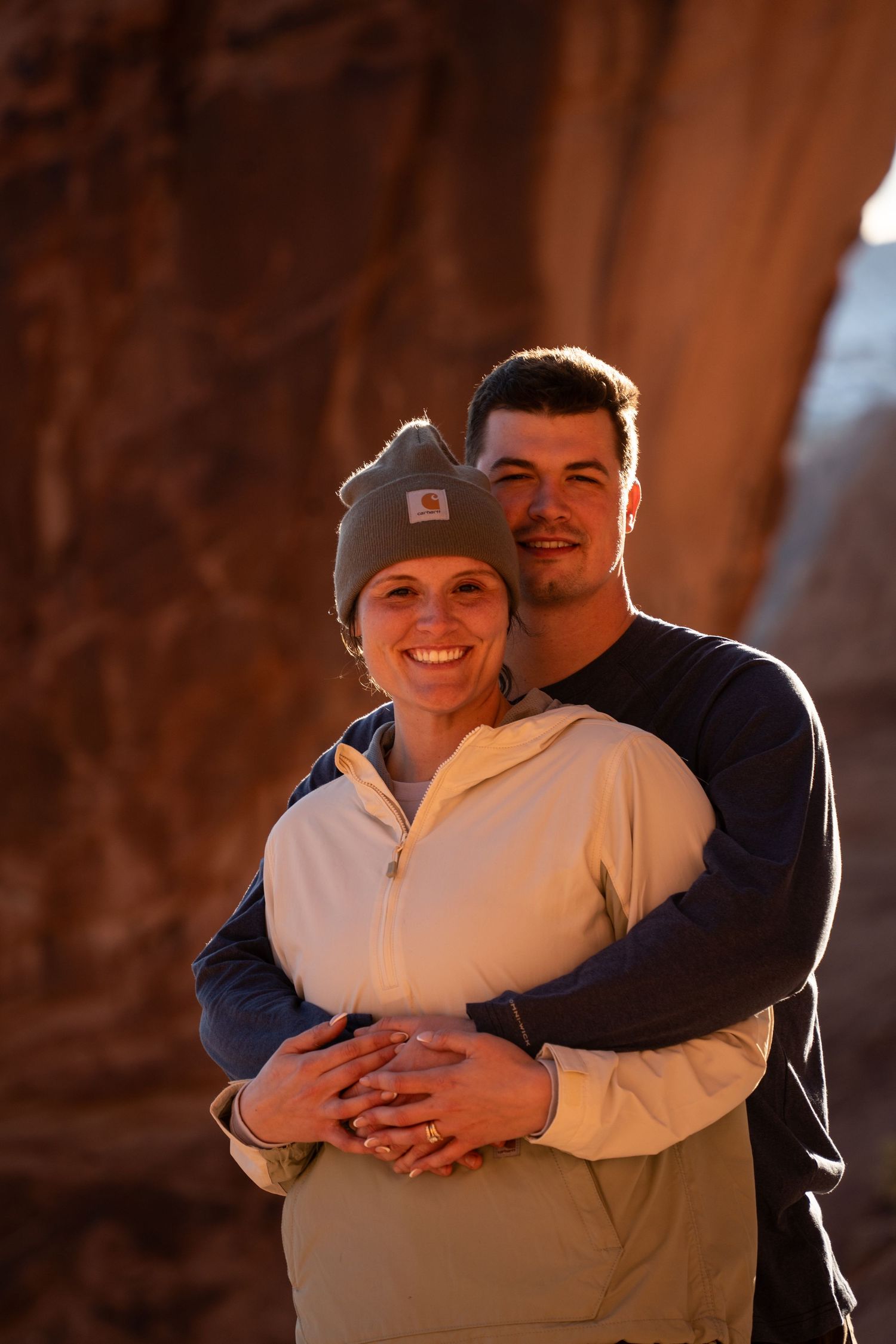 Couple embracing and smiling in a warm red rock canyon after adventure proposal, woman wearing a Carhartt beanie and fleece jacket.