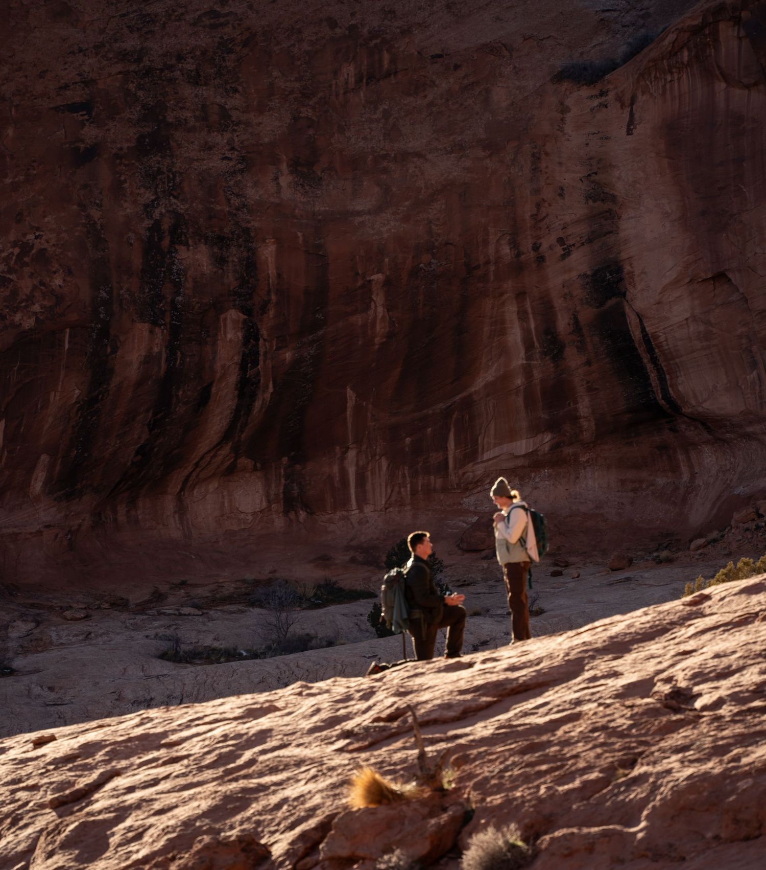 Two hikers stand on red rock formations before a towering sandstone canyon wall in a desert landscape.