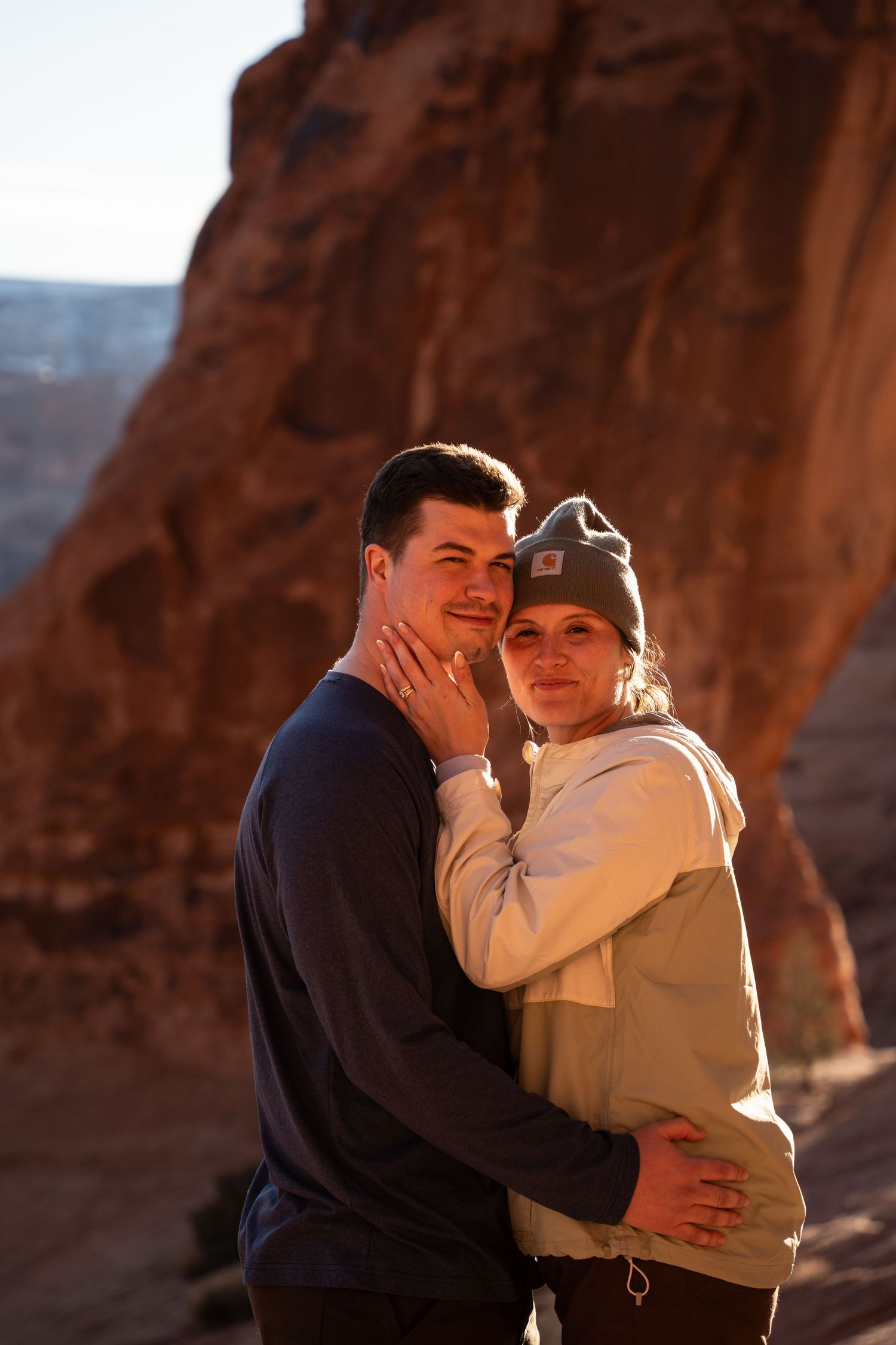 Couple embracing at golden hour in a red rock canyon moab utah proposal, woman in beanie resting hand on man's cheek.