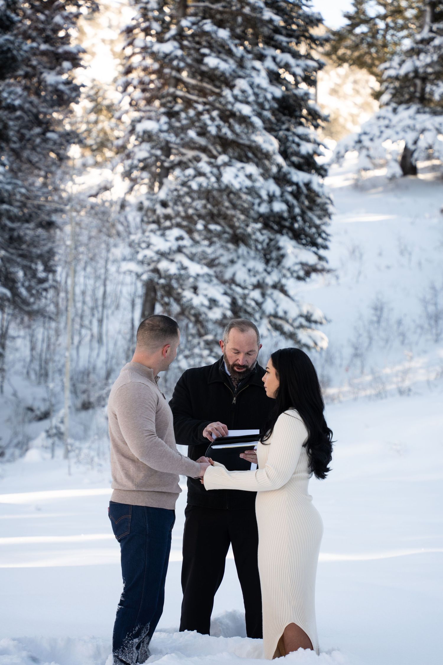 Couple exchanging vows in snowy winter outdoor ceremony with officiant, surrounded by snow-covered pine trees in Salt Lake Utah.