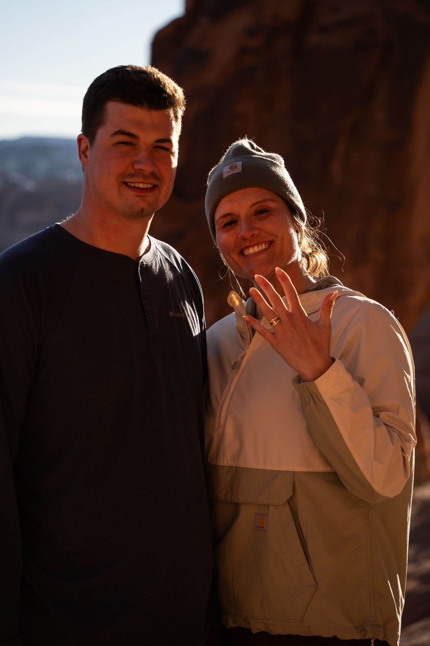 Happy couple smiling outdoors showing off engagement ring at golden hour near red rock formations in moab utah