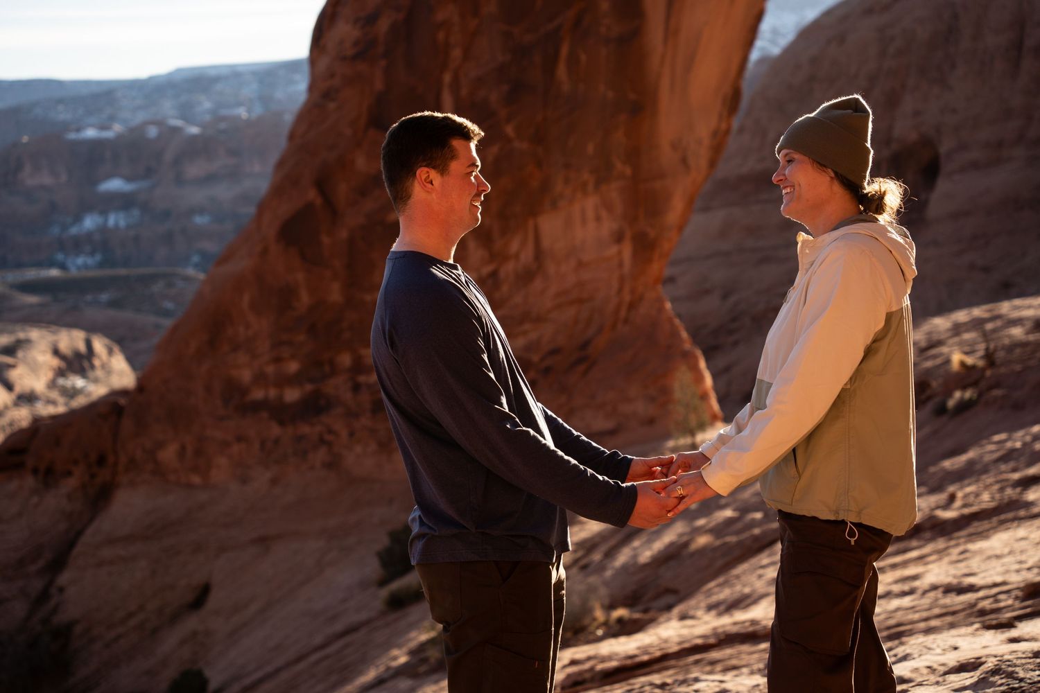 Couple holding hands facing each other on a red rock canyon overlook in Moab, Utah desert landscape.