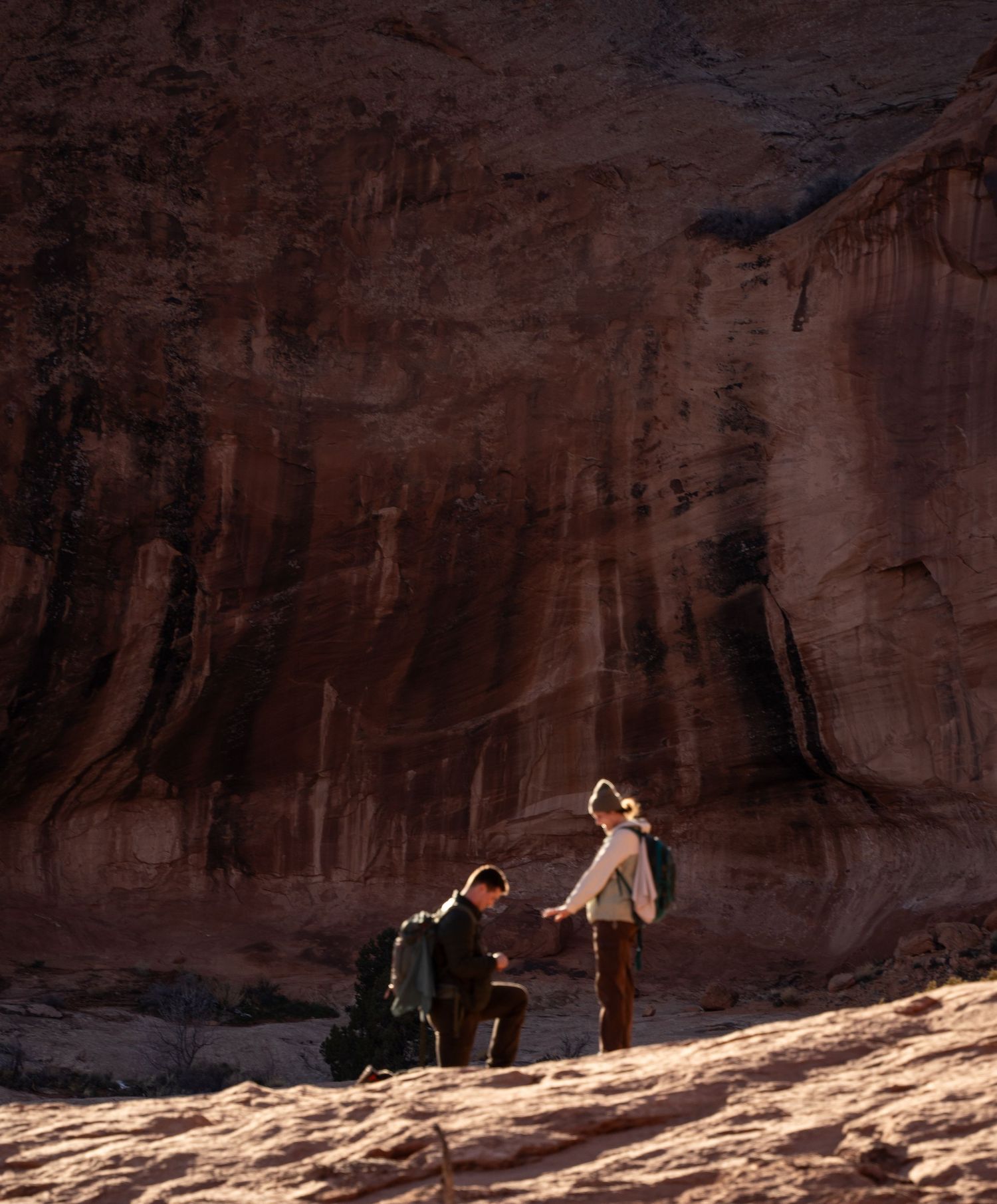 proposal at the base of a towering red sandstone canyon wall in a dramatic desert landscape.