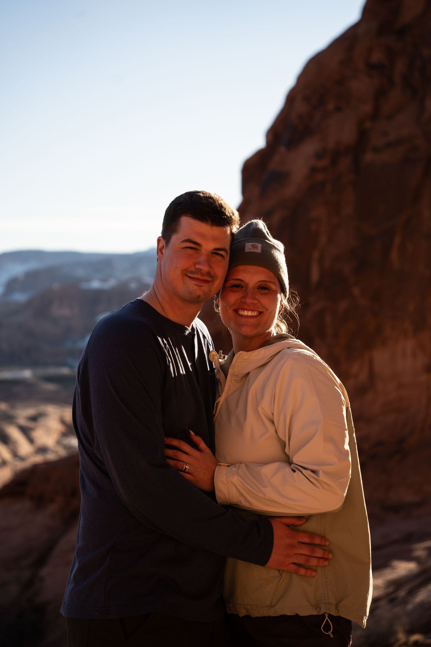 A couple embraces and smiles at a scenic desert canyon overlook with red rock formations in moab utah at golden hour.