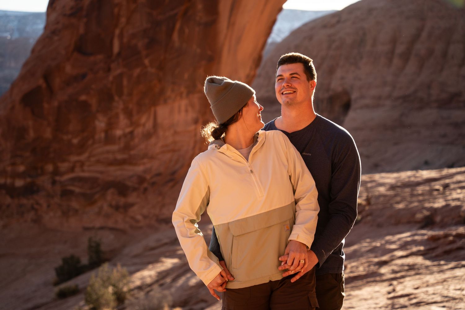 Couple smiling and embracing at sunset in a red rock canyon landscape, wearing casual outdoor clothing.