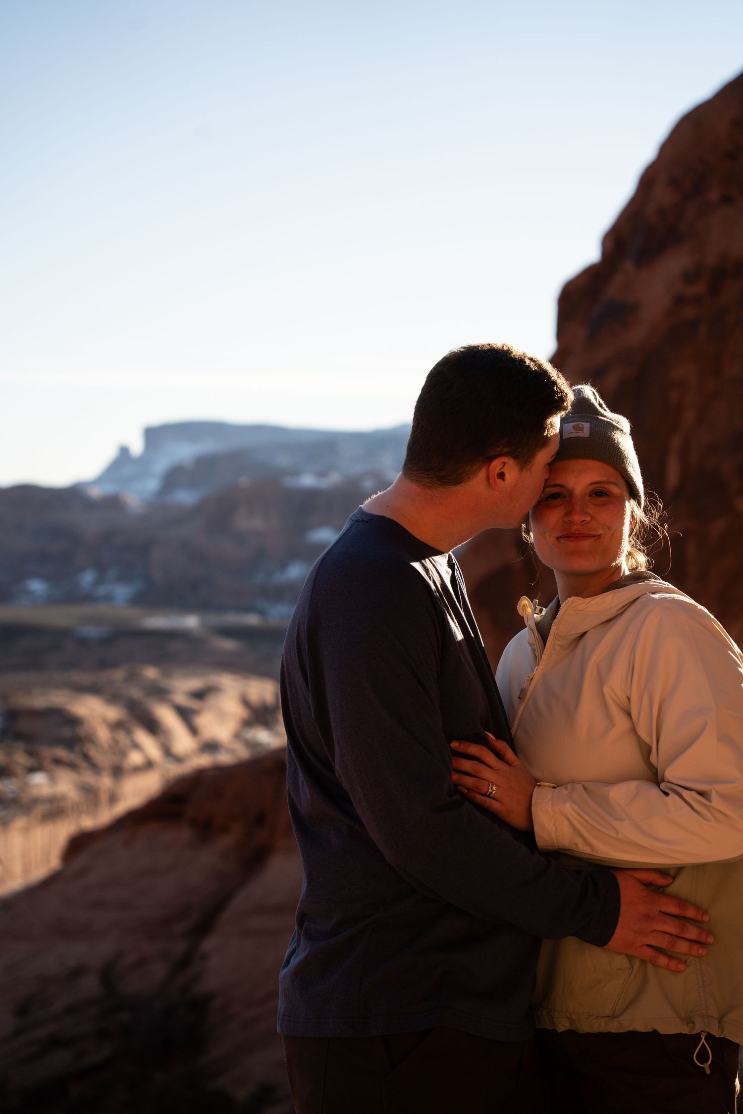 Couple embracing on rocky desert overlook at golden hour with red rock formations and blue sky backdrop.