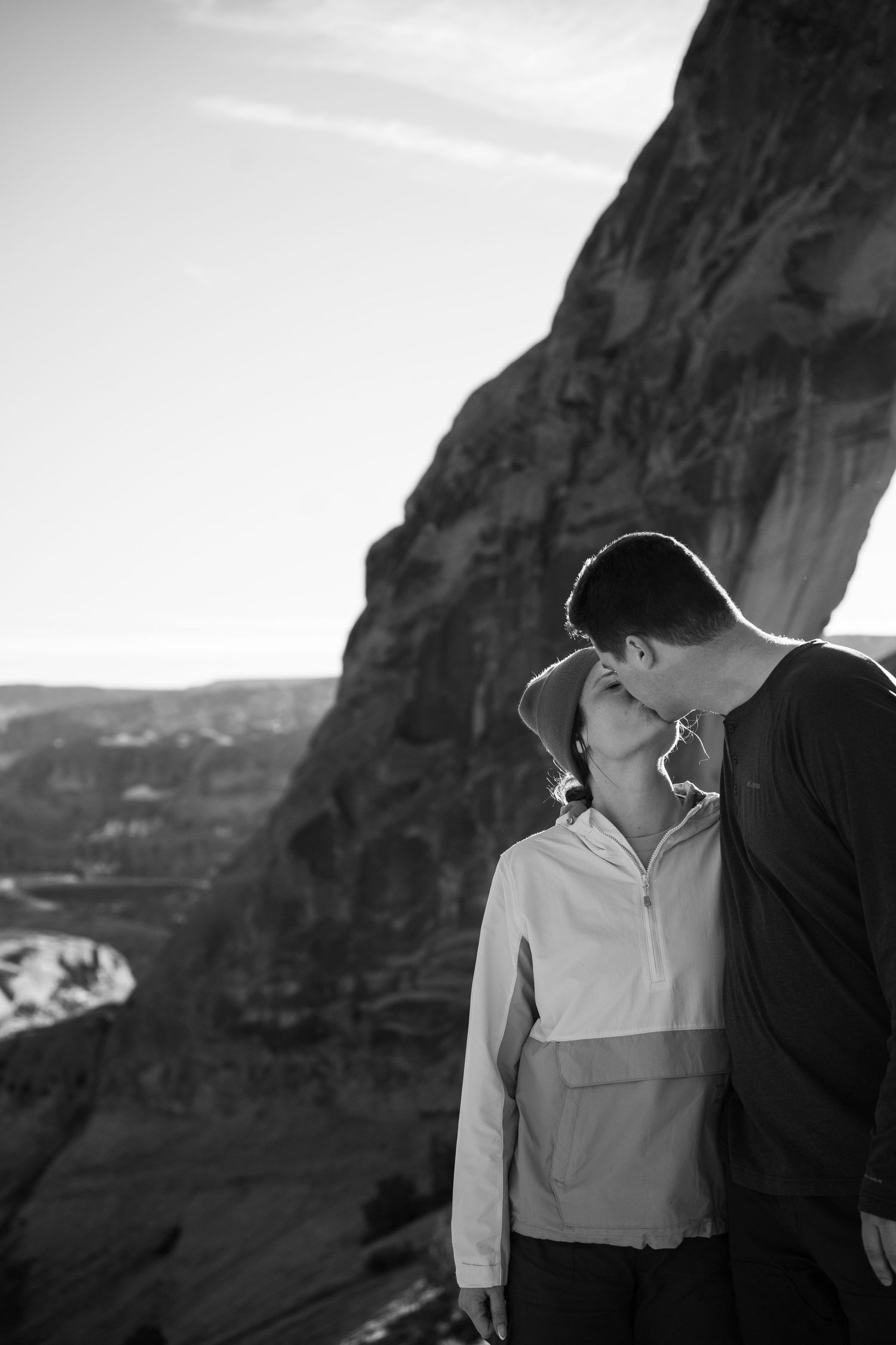 Black and white photo of a newly engaged couple kissing in front of dramatic rock formations and canyon landscape.