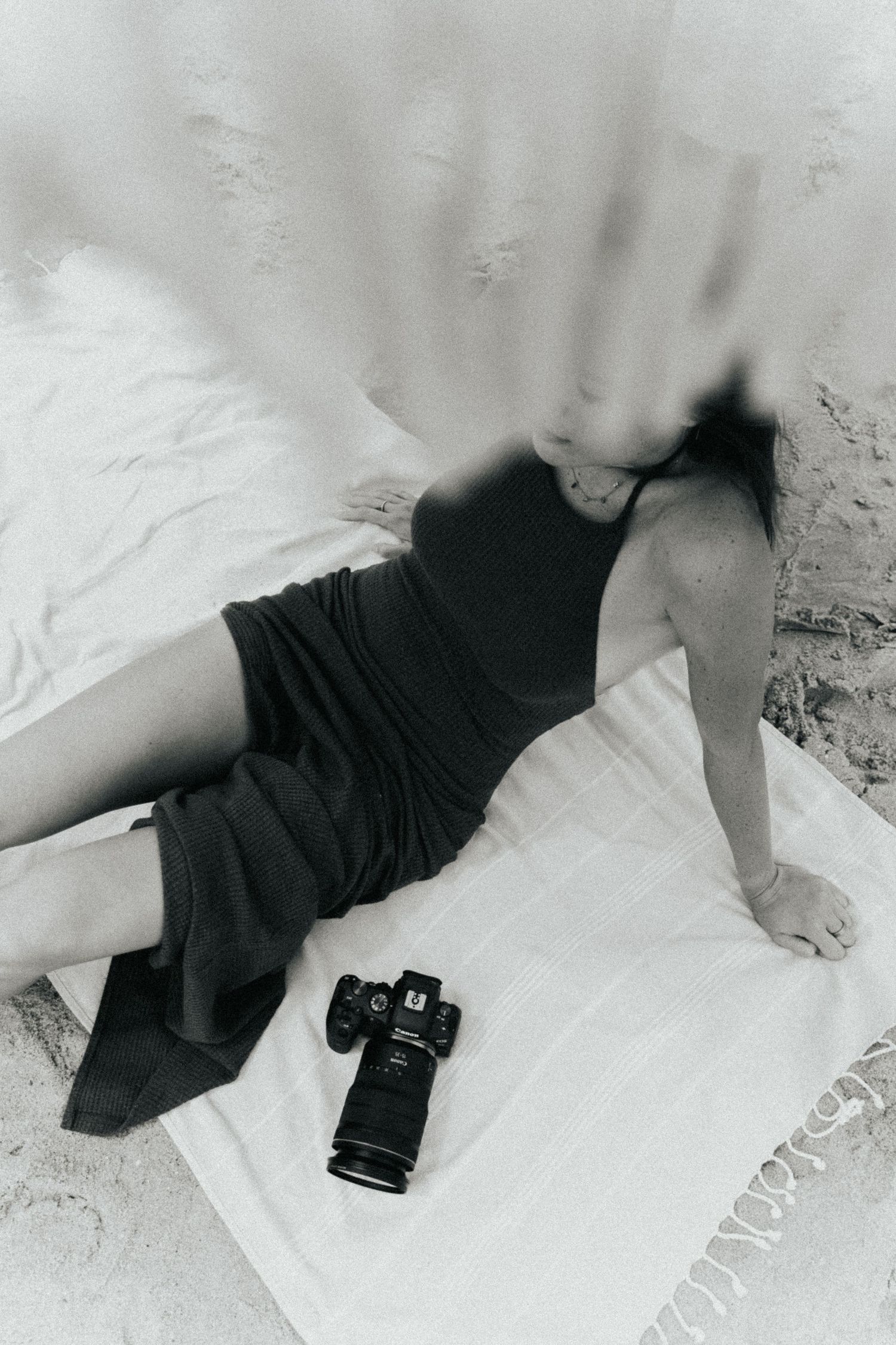 Black and white photo of a woman in a dark dress lying on a beach towel with a DSLR camera beside her.