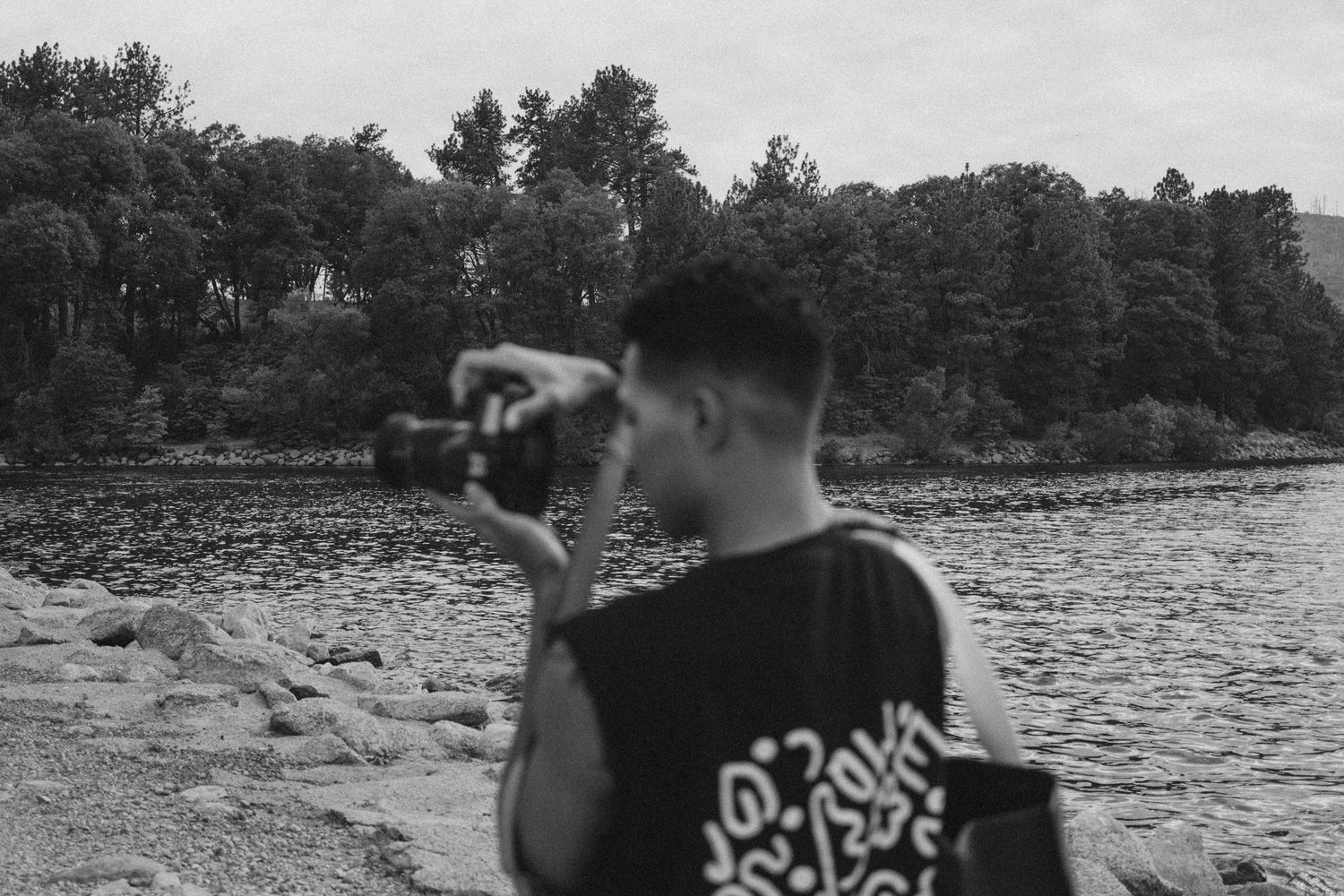 Black and white photo of a young man photographing a rocky riverbank with dense trees in the background.