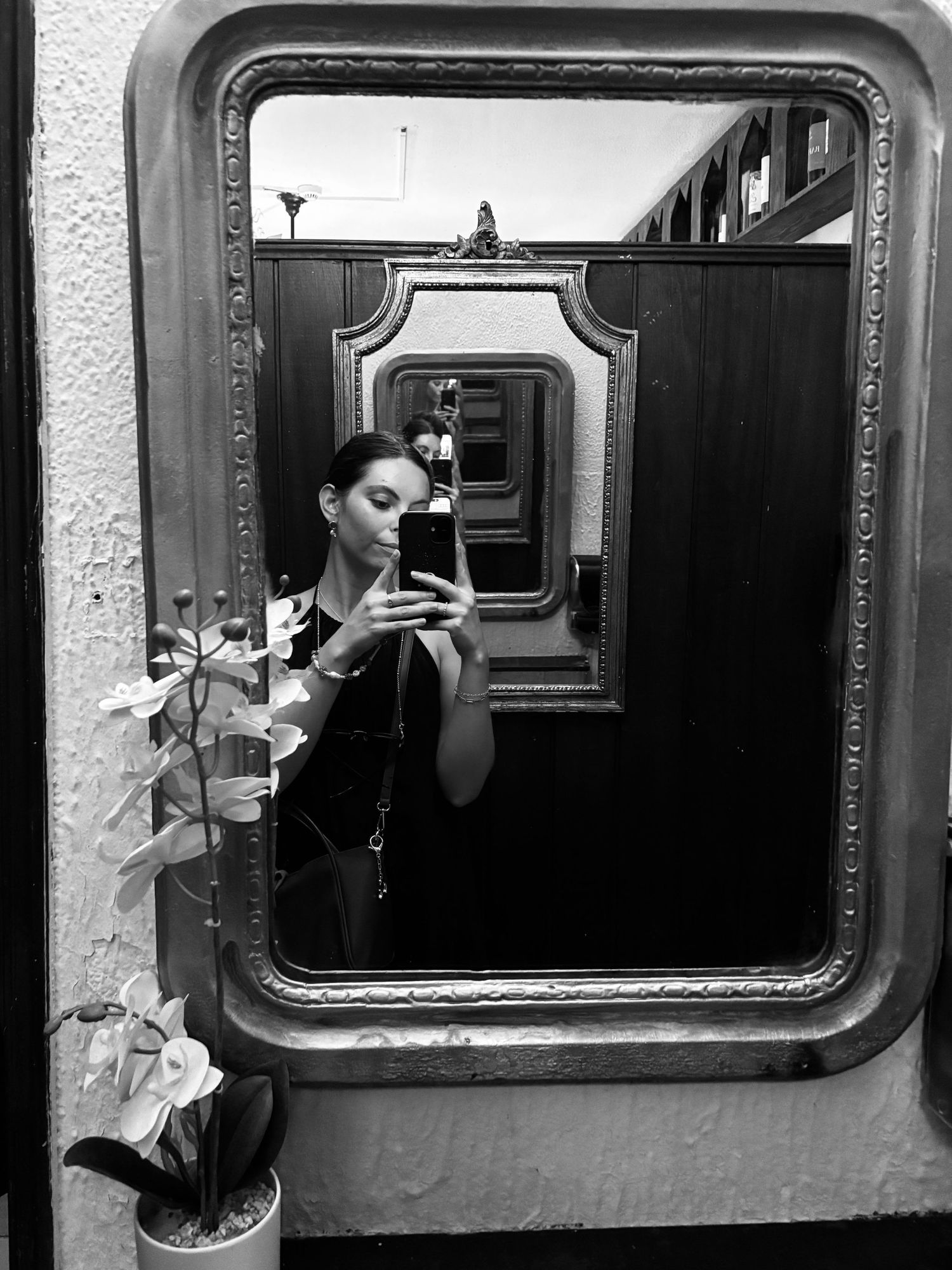 Black and white mirror selfie of a young woman holding a phone in an ornate gold-framed mirror with a flower vase nearby.