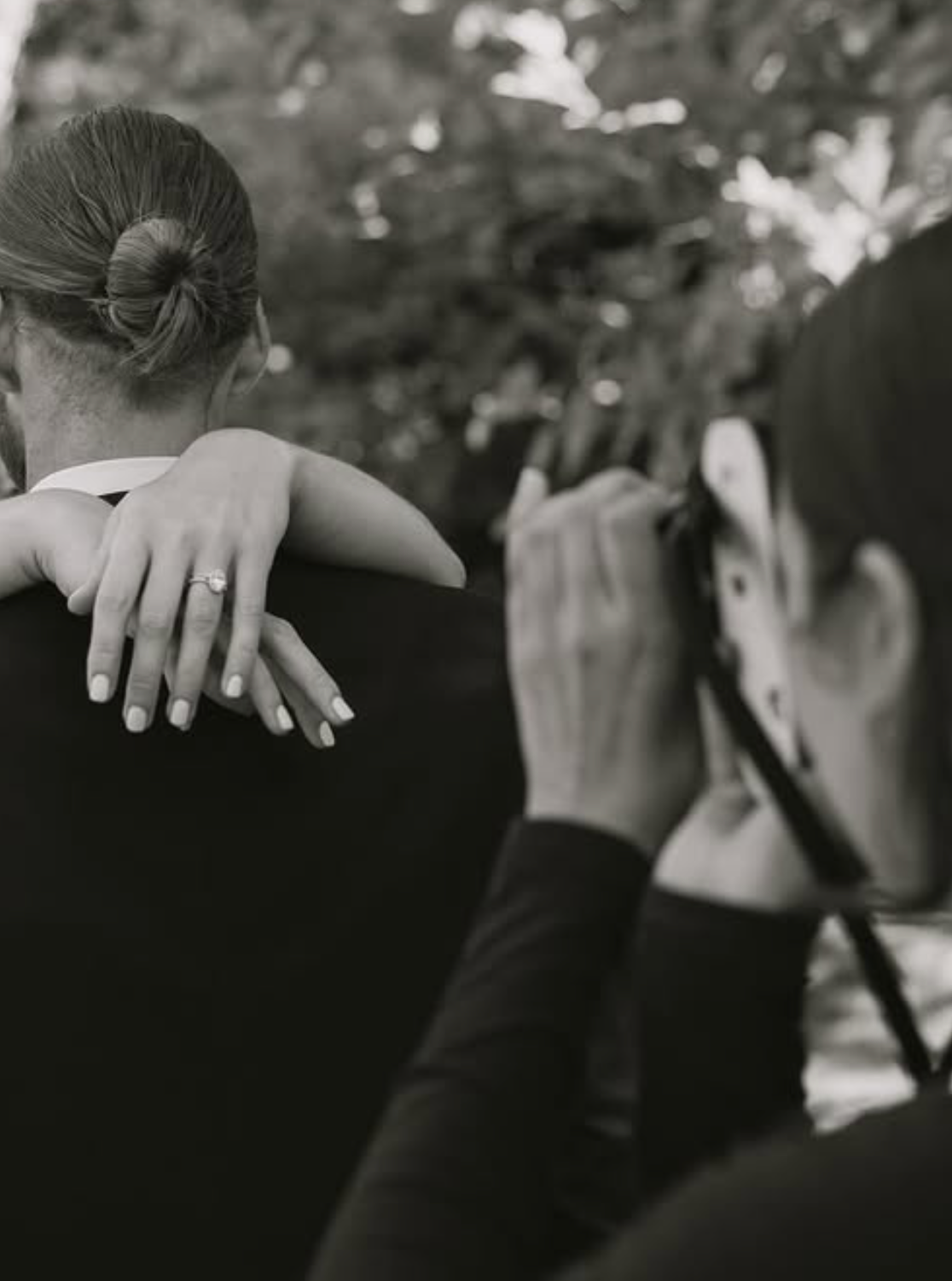 Black and white photo of bride with engagement ring on groom's shoulder while photographer captures the moment.
