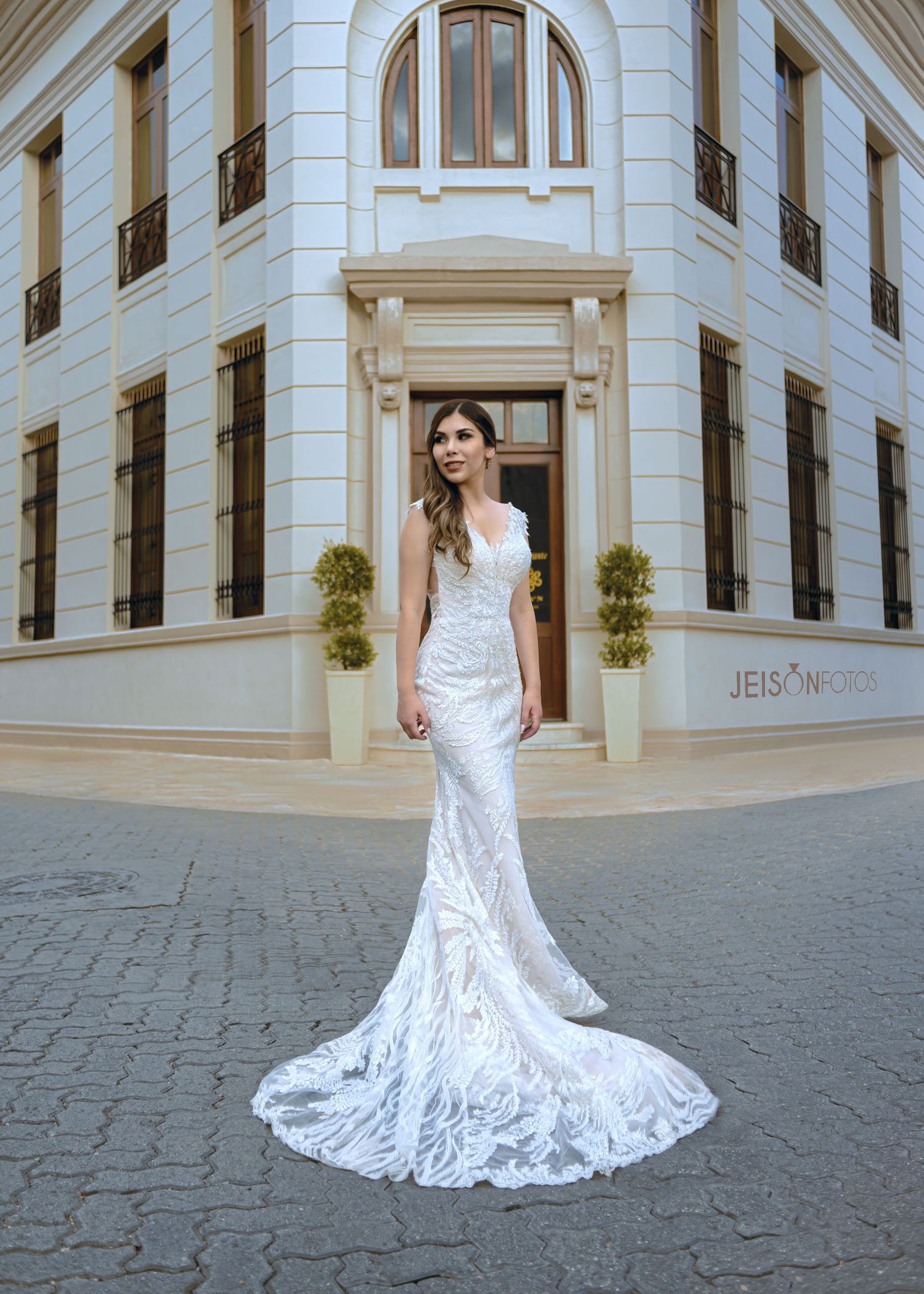 Una novia con un vestido de boda de encaje blanco, fluido y con una larga cola, posa en un elegante pasillo de mármol.
