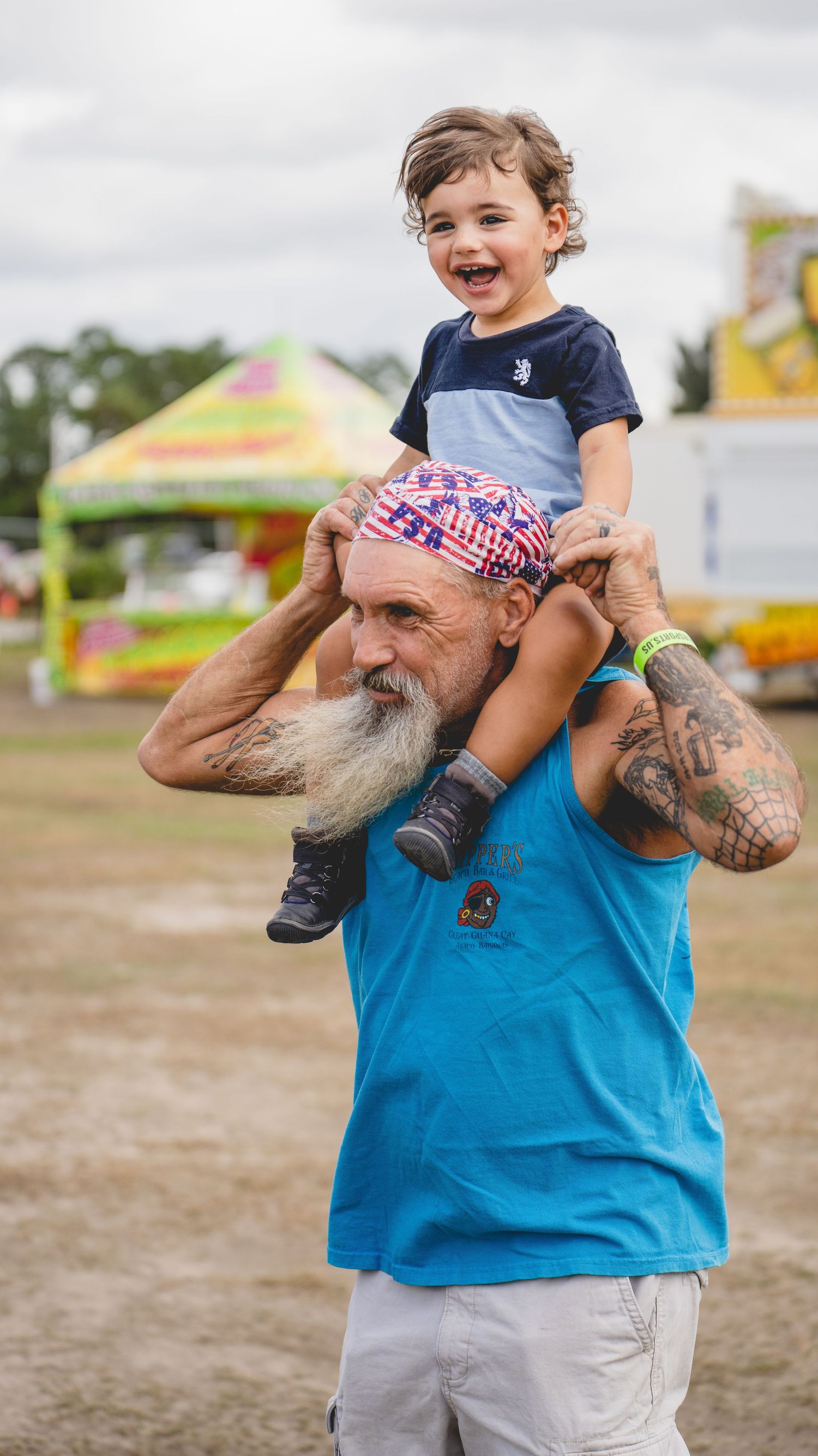 A bearded man in a blue tank top carries a young child on his shoulders at an outdoor event.