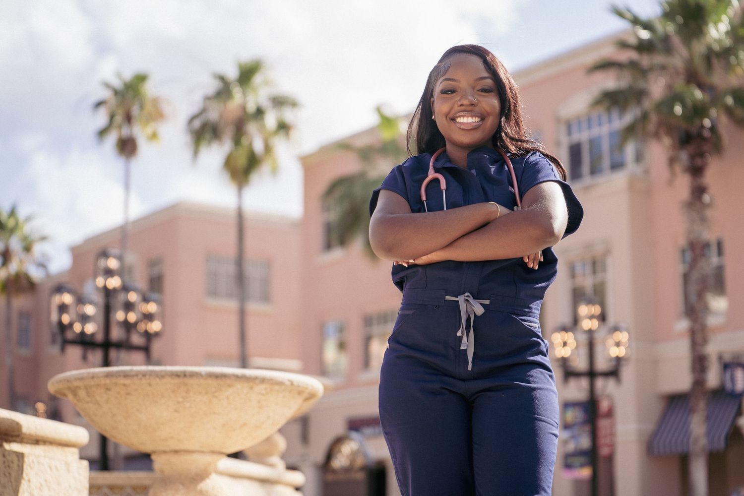 A person in a navy blue outfit poses in front of a pink building with palm trees on a sunny day.