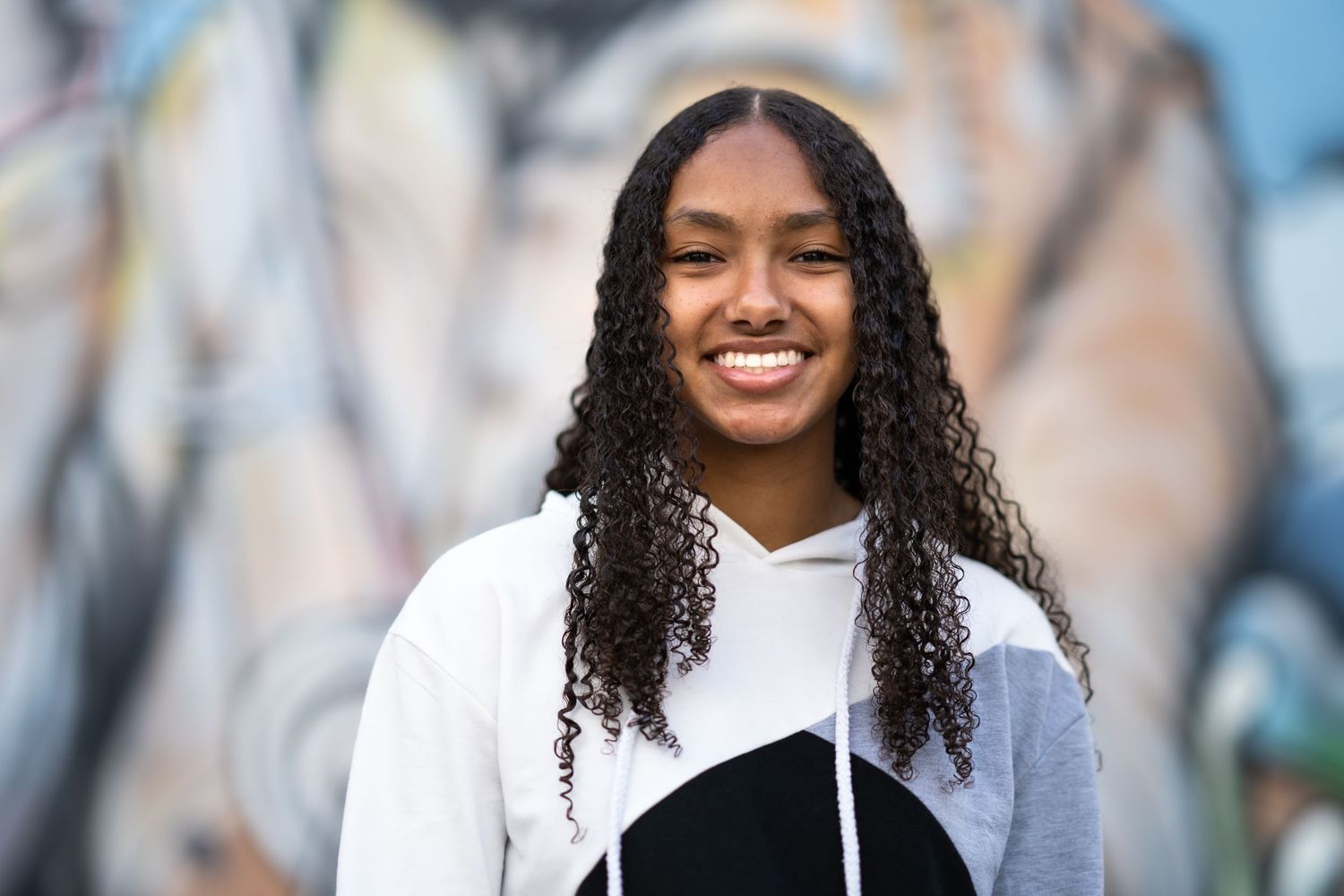 A person with long curly dark hair and a bright smile wearing a black and white hoodie stands against a blurred wall.