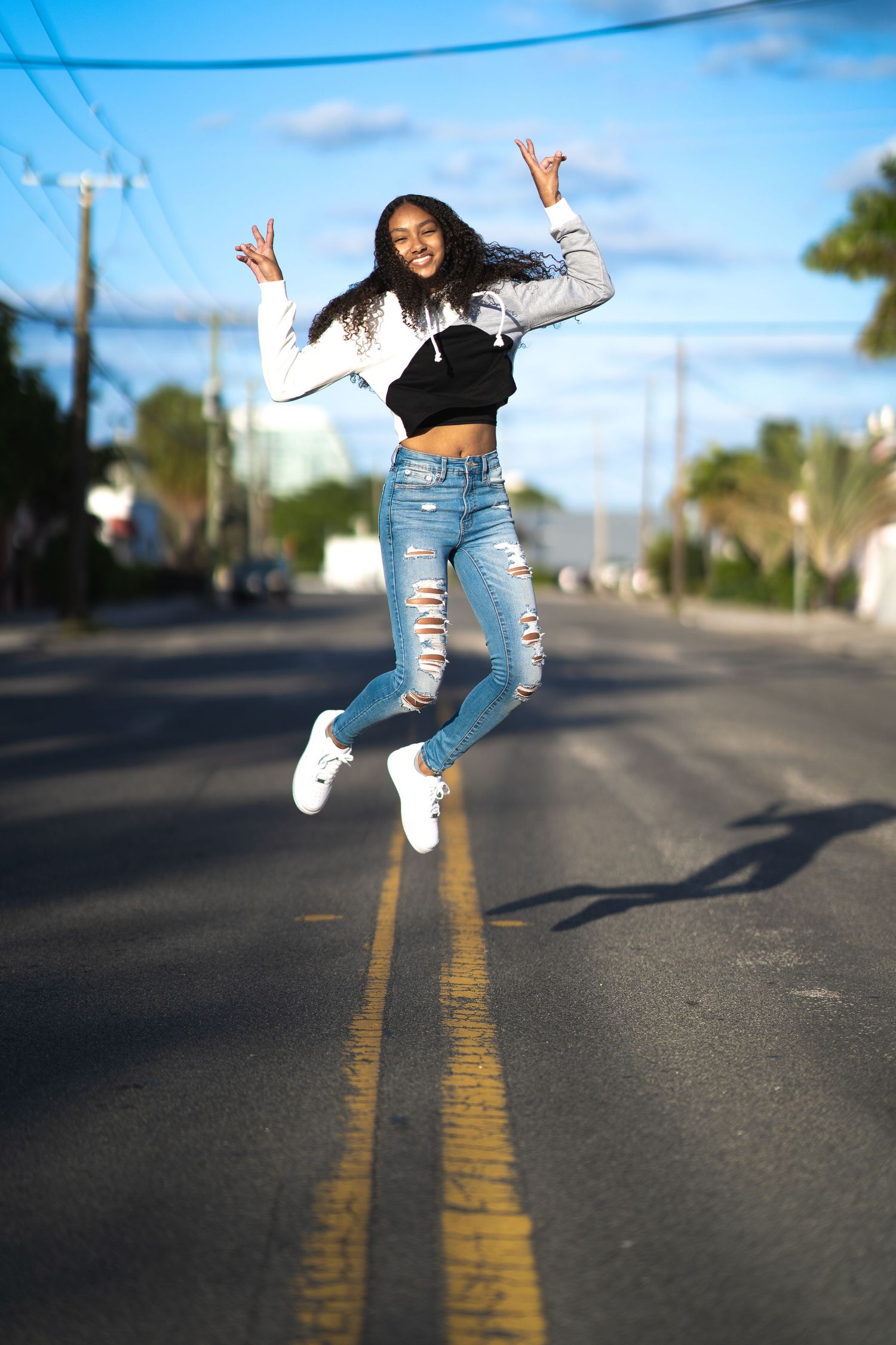 Energetic young woman in ripped jeans and white crop top jumps playfully over double yellow lines on an empty street.