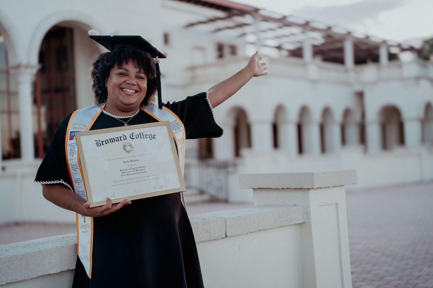 Graduate holds up diploma with excitement while standing on white steps during outdoor graduation ceremony.