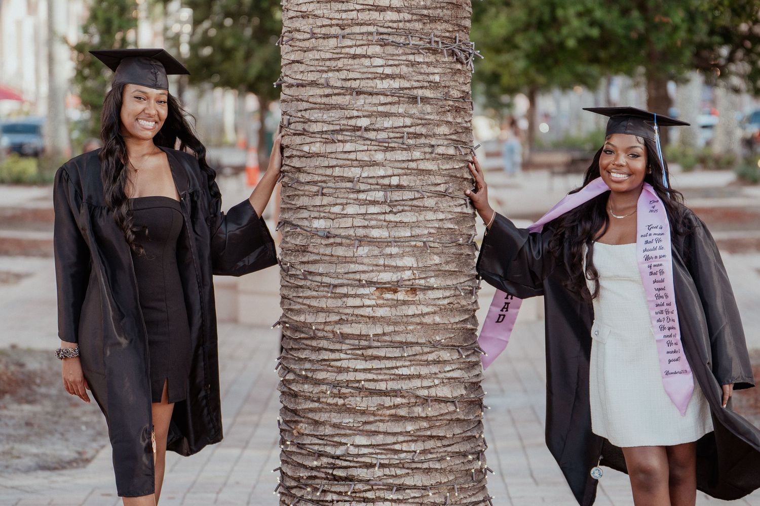 Two graduates in black caps and gowns pose on opposite sides of a palm tree trunk during graduation celebrations.