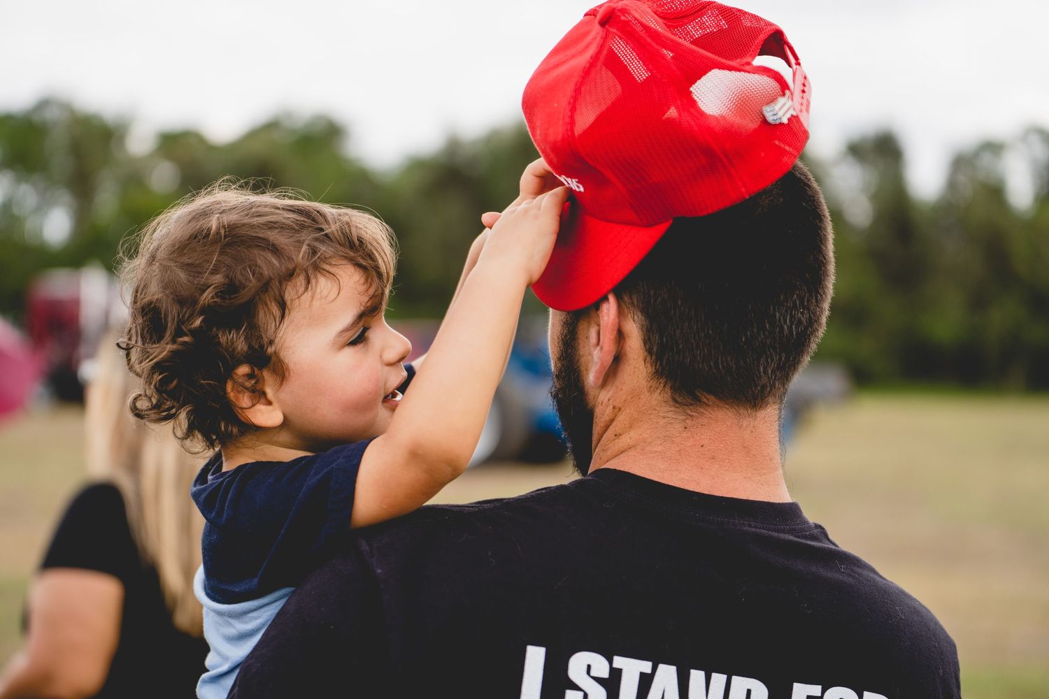 Child placing red cap on person wearing black t-shirt in outdoor setting.