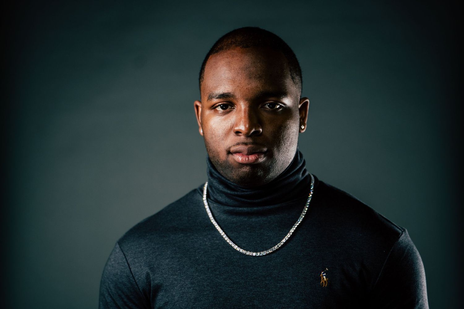 Dramatic headshot featuring person in black turtleneck with silver necklace against moody backdrop.