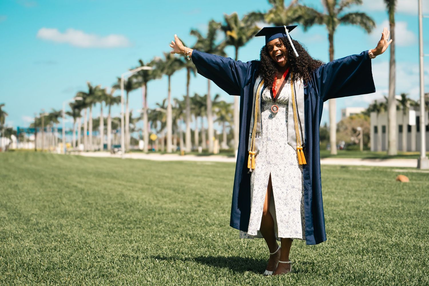 A person celebrating in a blue graduation robe stands on grass with palm trees in the background.