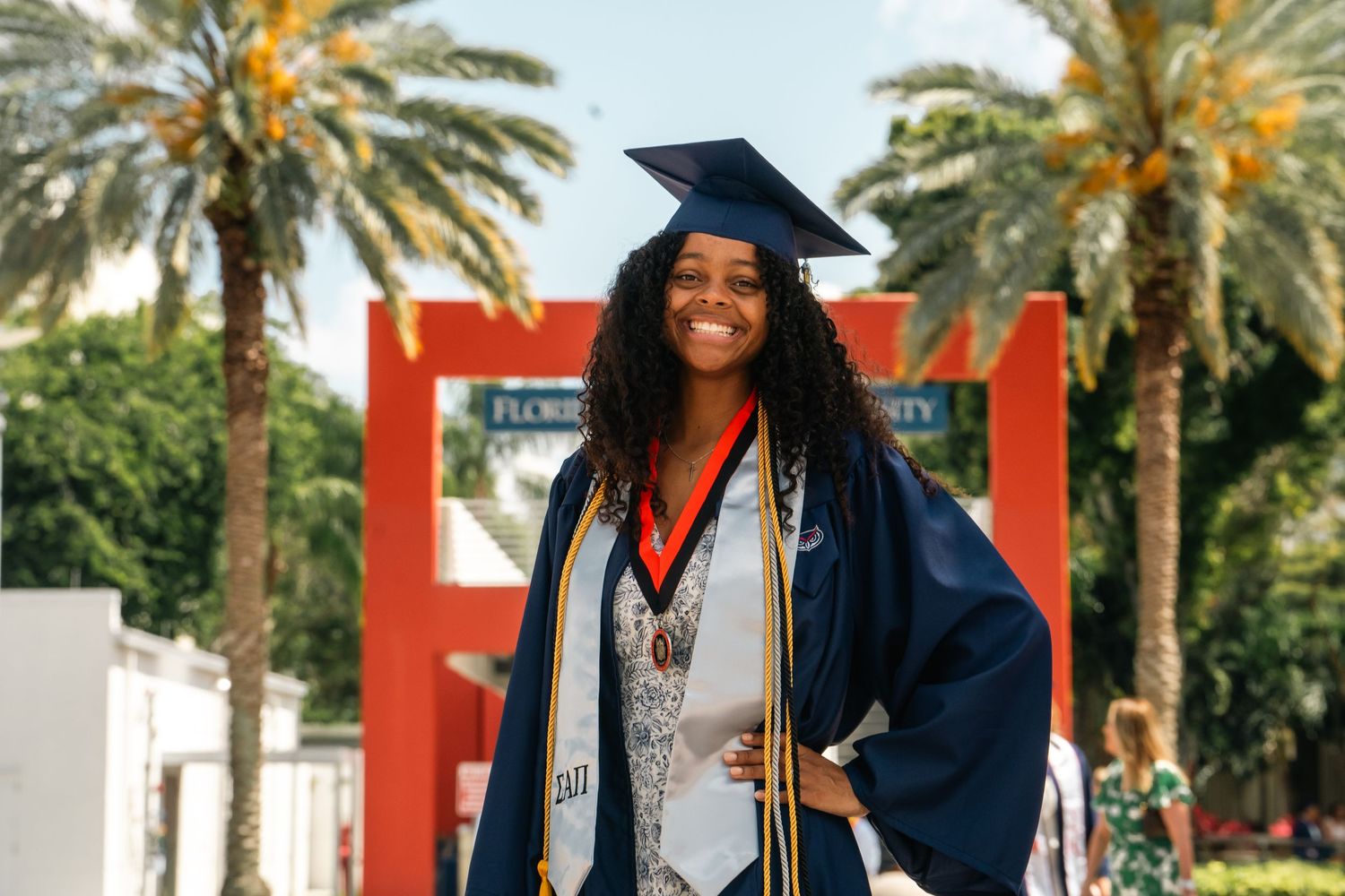 Graduate in blue cap and gown posing in front of palm trees and red architectural elements.