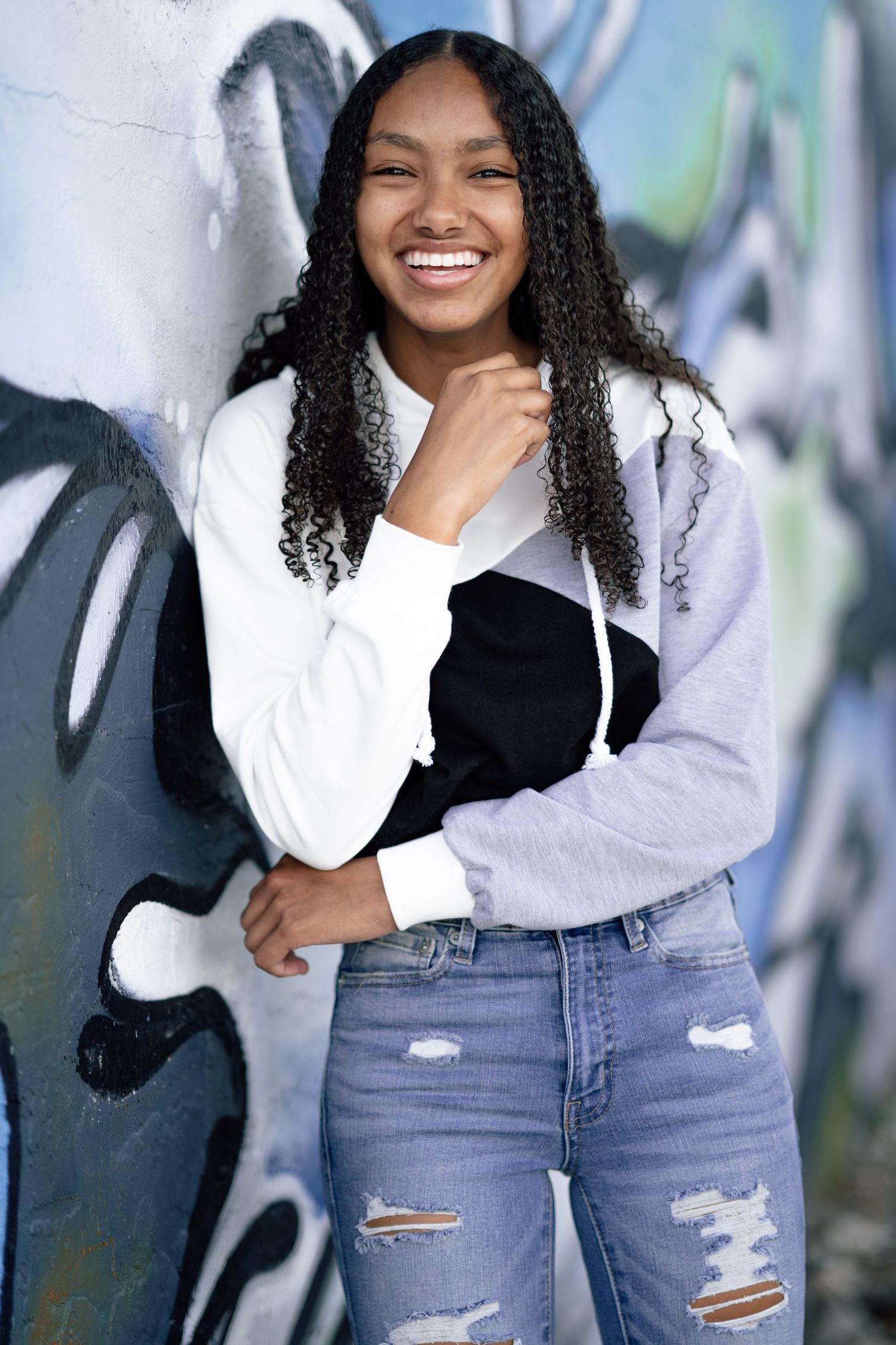 Young woman in gray hoodie and ripped jeans poses against graffiti wall with natural curly hair.