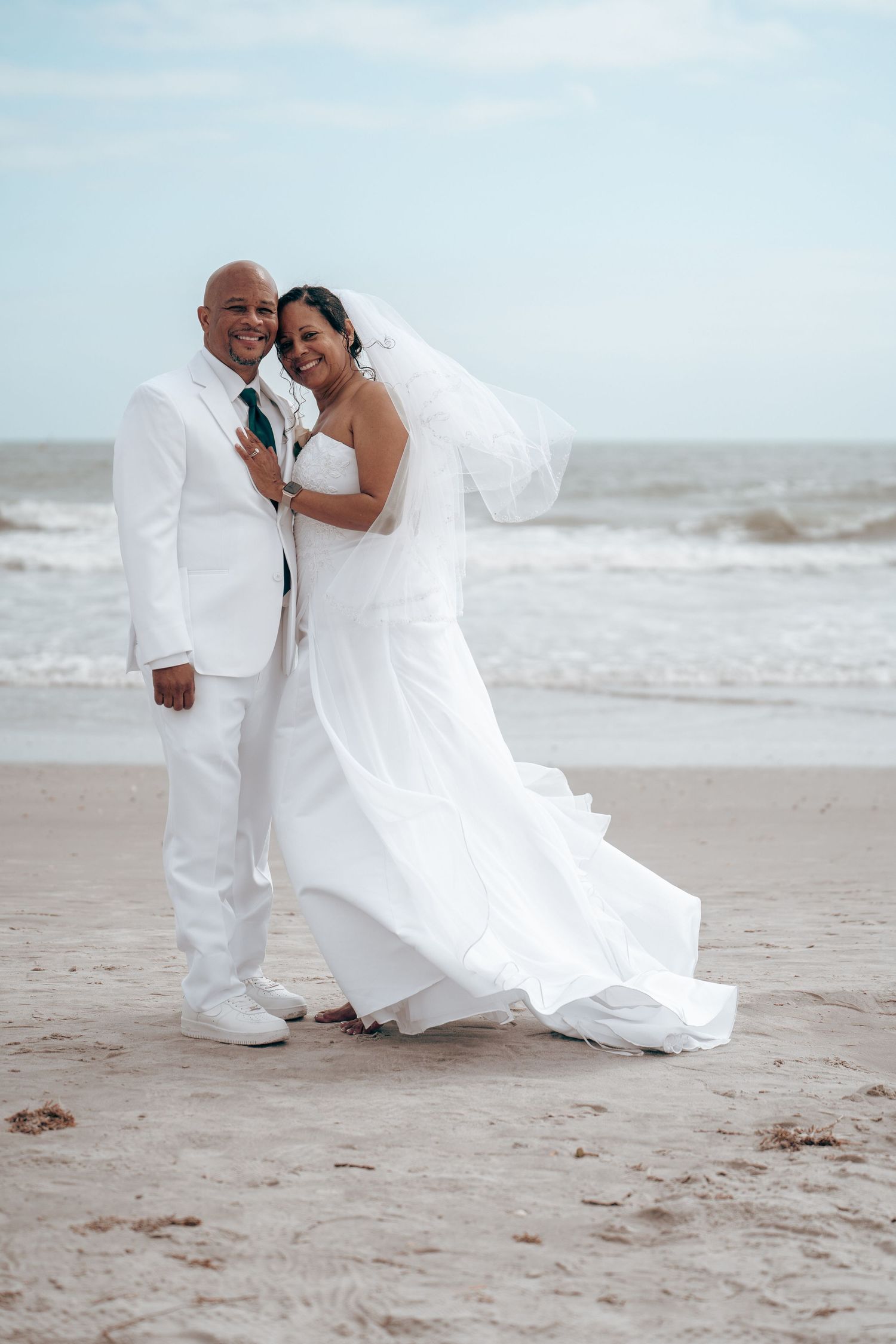 Romantic beach wedding photo with couple in white attire as wind catches the veil.