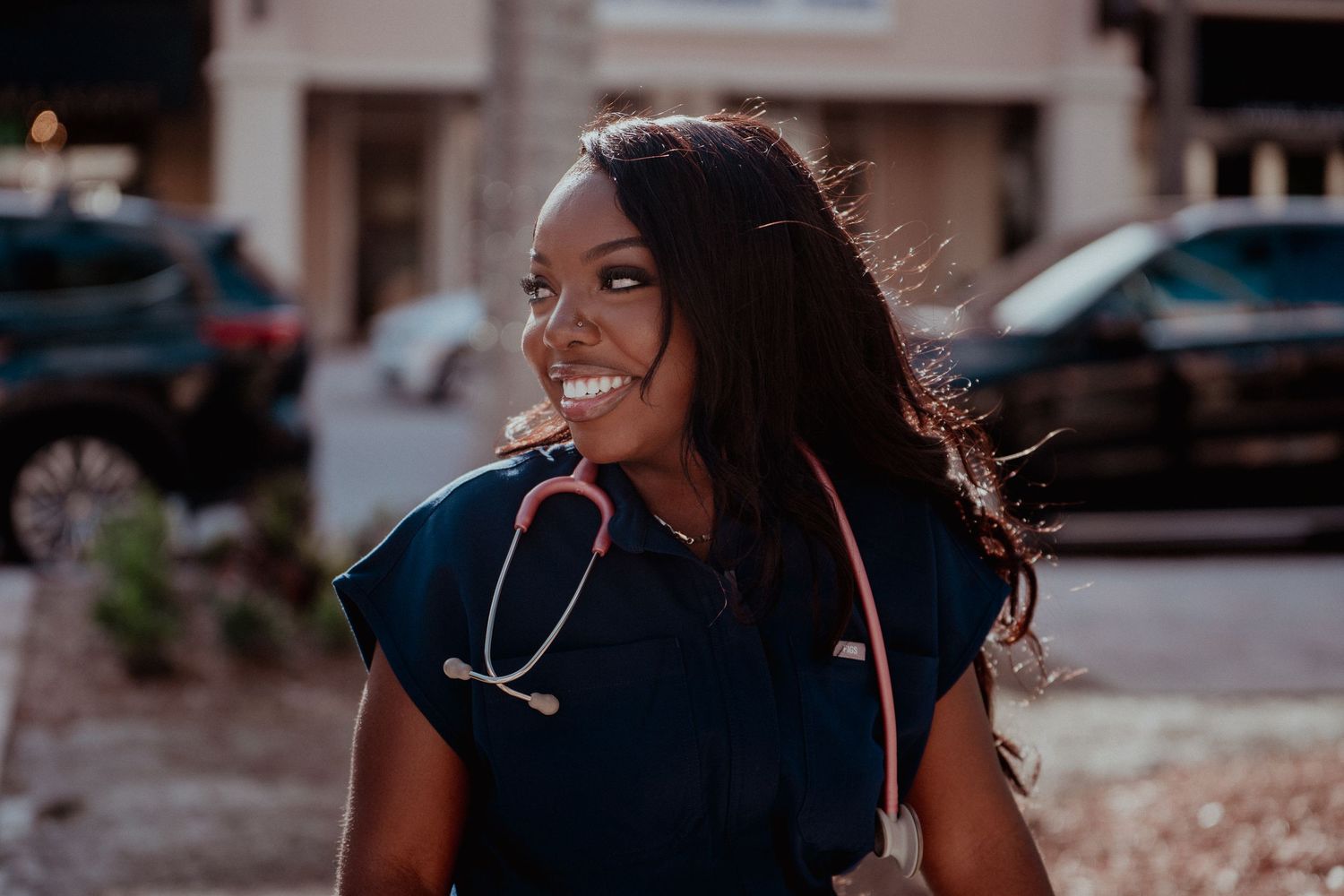 A medical professional in navy scrubs with a stethoscope smiles while standing outside in warm sunset lighting.