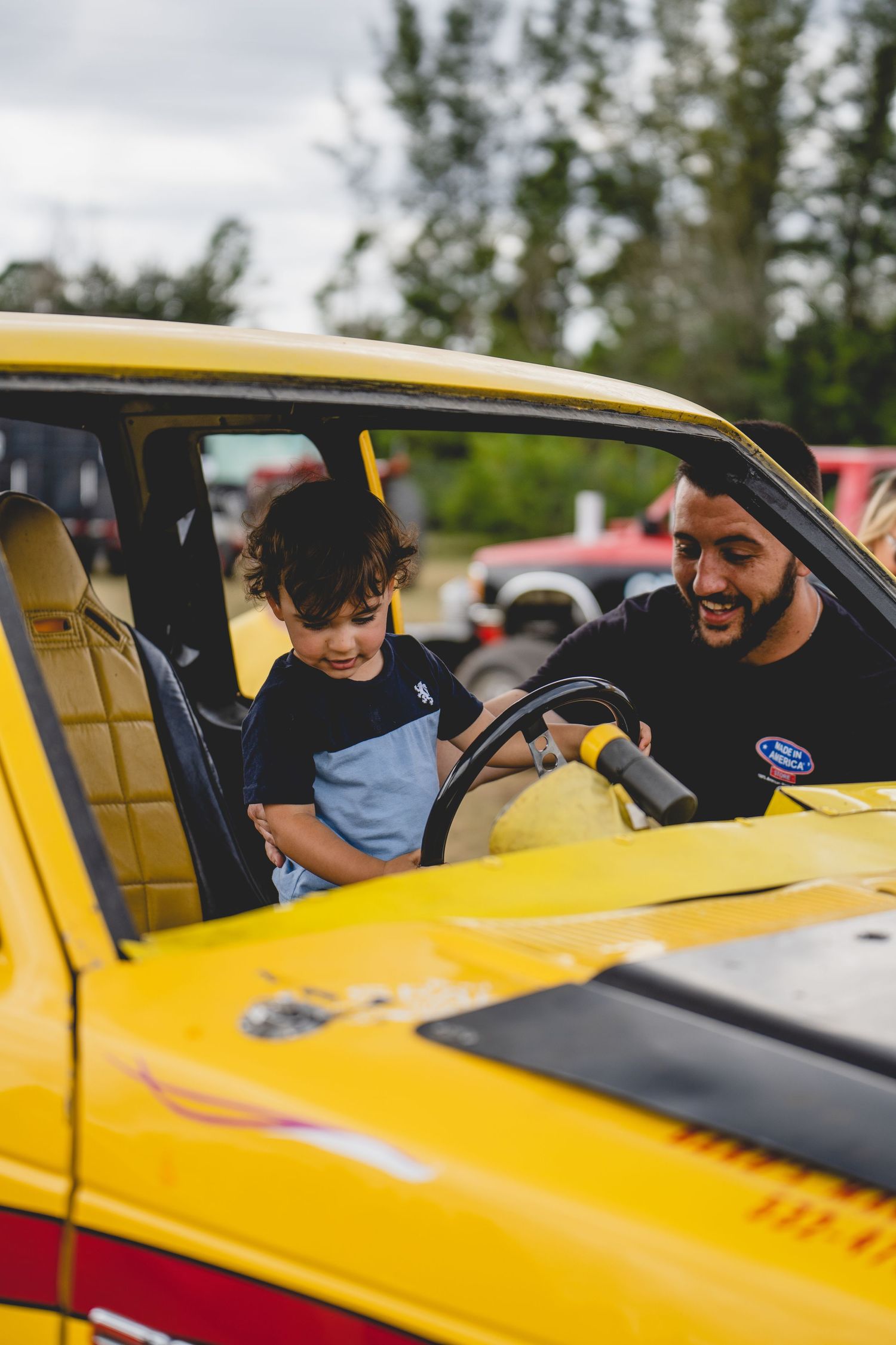 A child explores the interior of a bright yellow classic car while an adult looks on smiling.