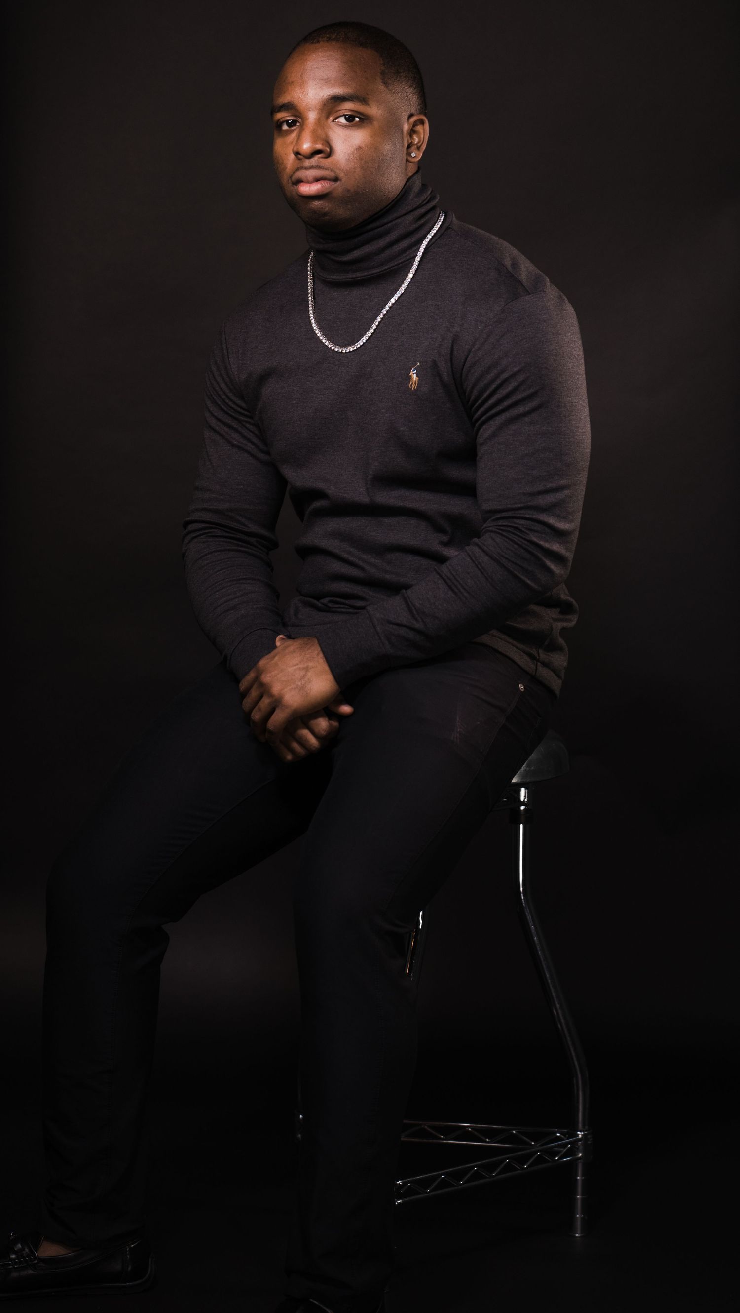 Professional studio portrait of person in black turtleneck with silver chain against dark background.