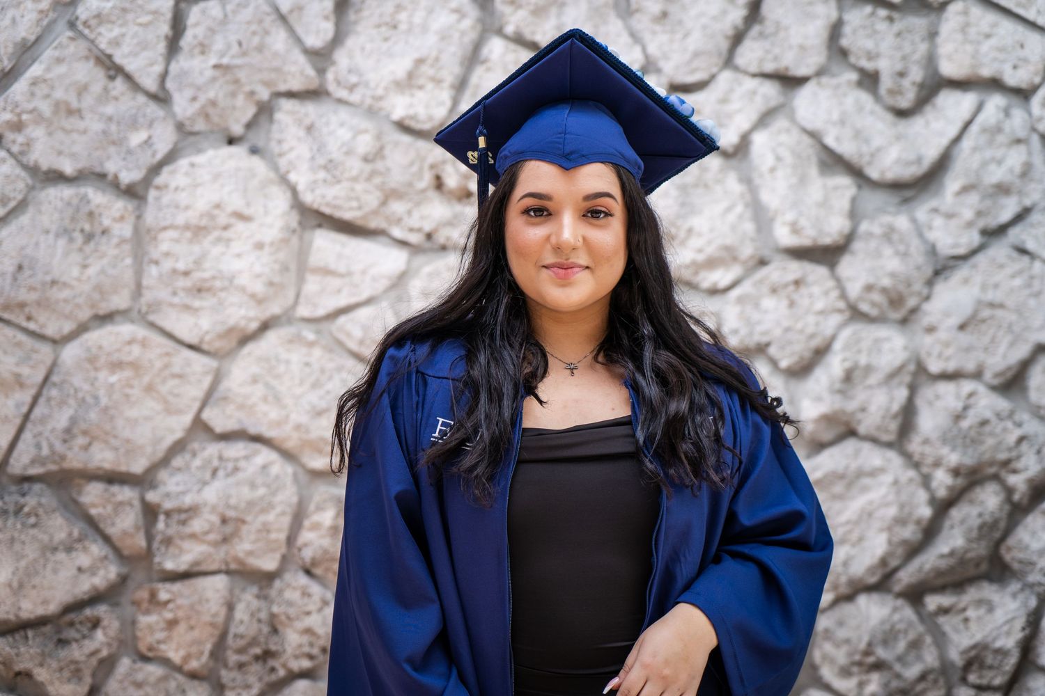 Graduate in blue academic regalia poses in front of a textured stone wall.