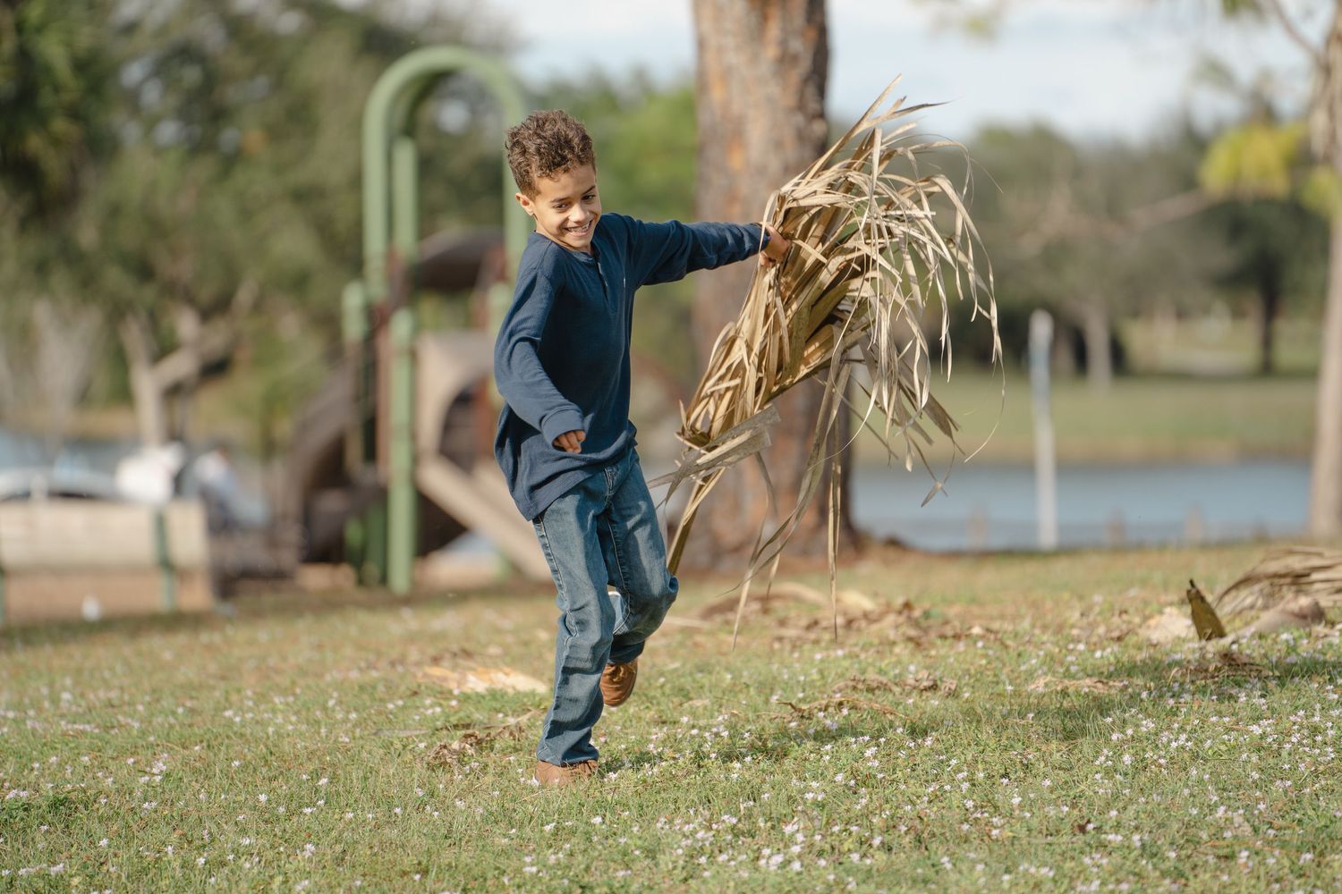 Child playing and jumping outdoors in a park with trees and water in the background.