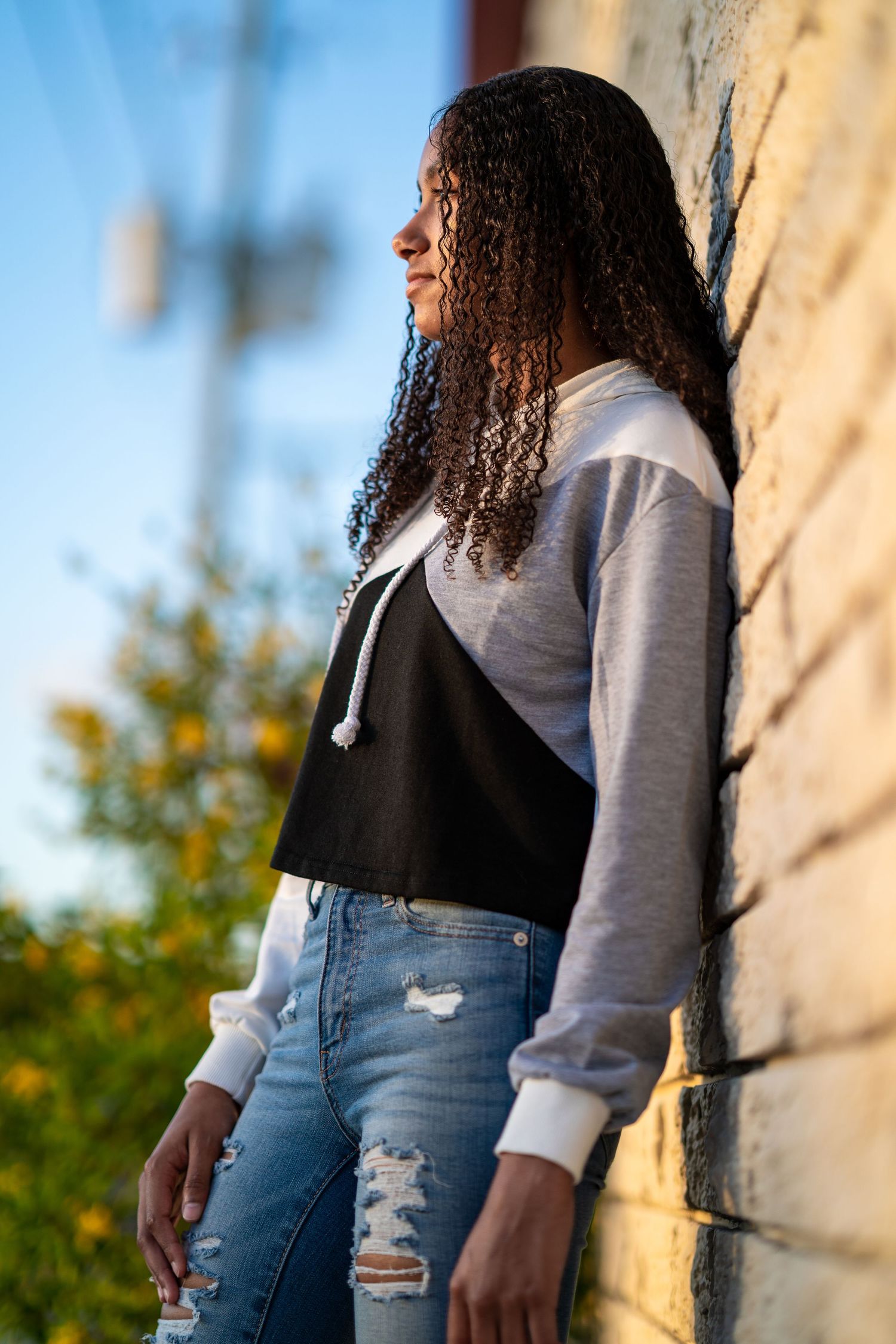 Side profile portrait of someone with curly hair wearing a gray sweatshirt, black top and distressed jeans against a wall.