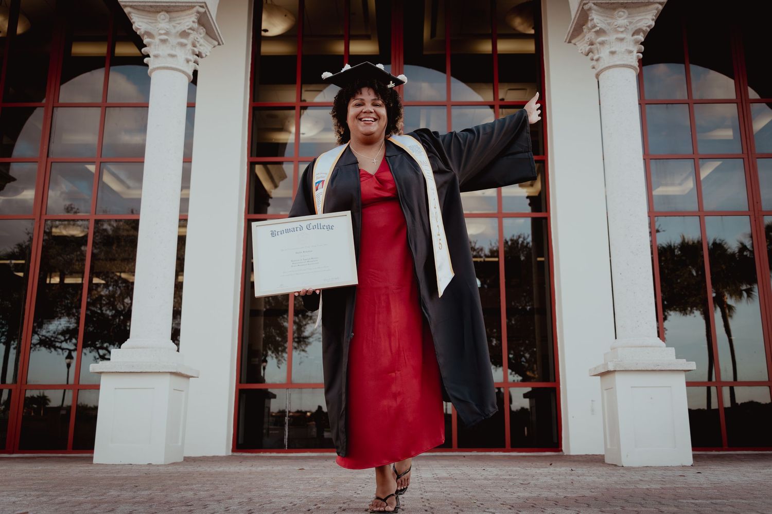 A person in a red dress and black jacket poses in front of white columns and glass windows.