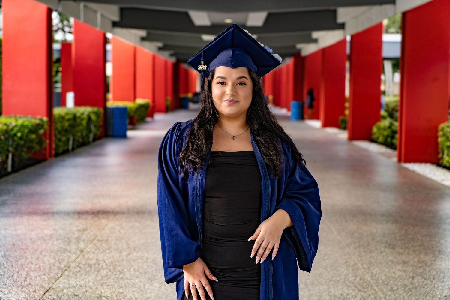 Graduate in blue cap and gown posing in a covered walkway with red columns.