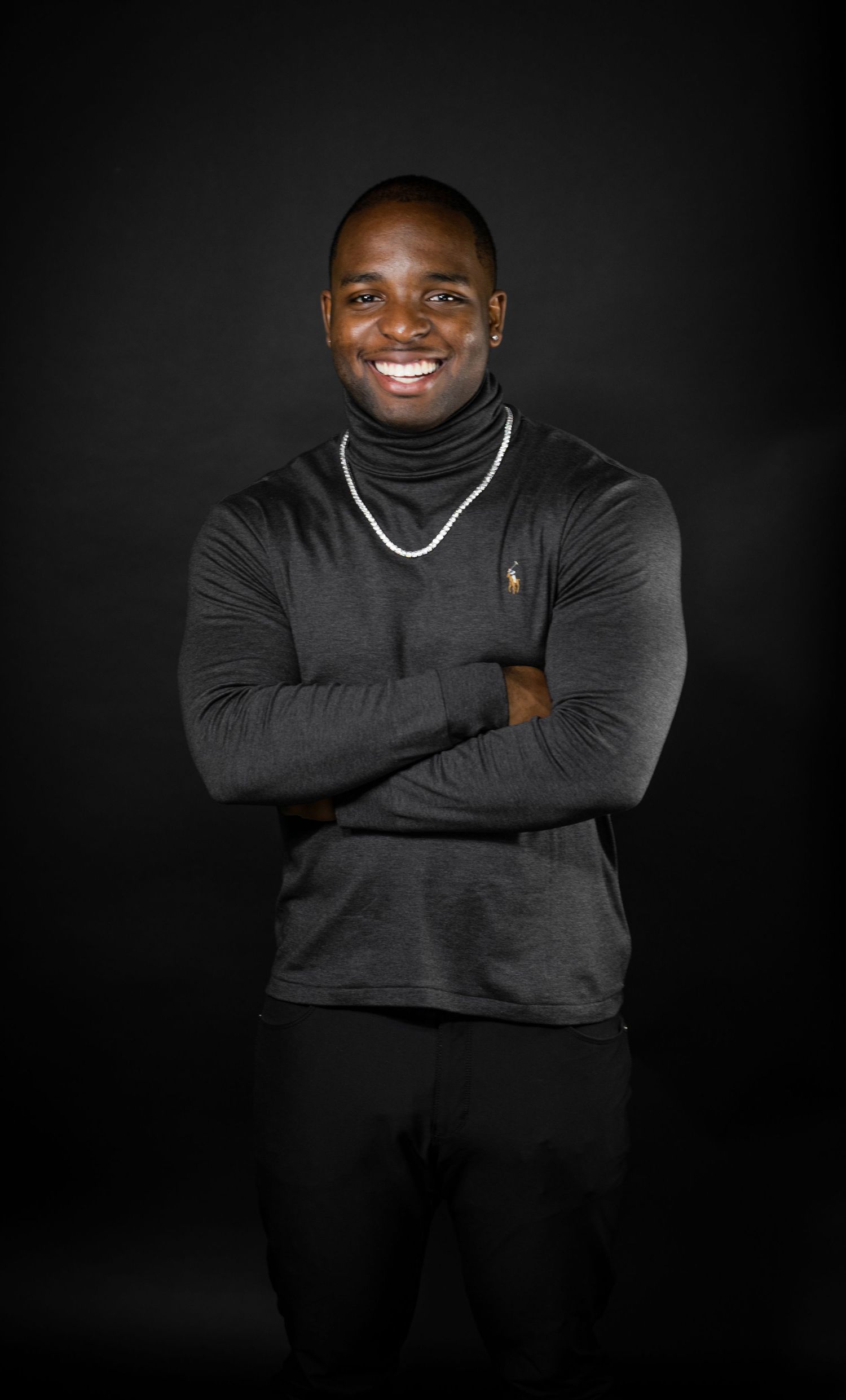 Professional portrait showing person in black turtleneck with silver chain smiling against dark backdrop.