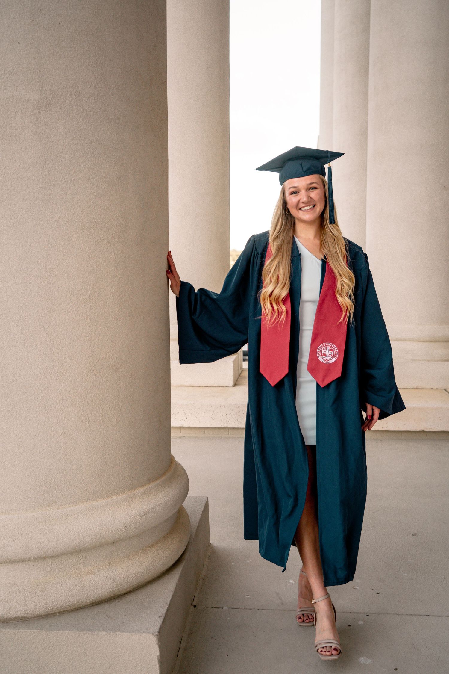 Smiling female graduate in navy cap and gown with red stole poses between white columns for graduation photos.