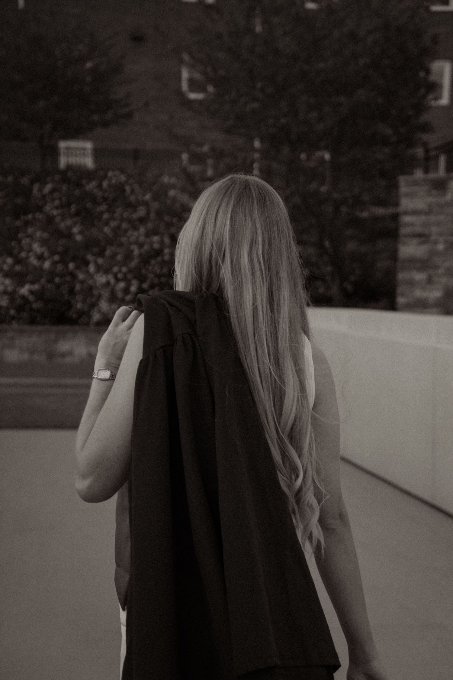 Black and white photo of a blonde woman seen from behind, wearing a black top outdoors near brick buildings.