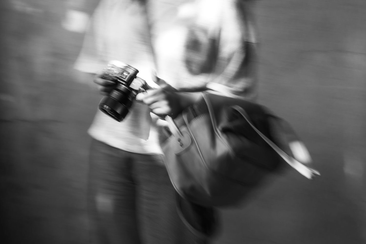 Blurred black and white photo of person holding camera and bag while walking.