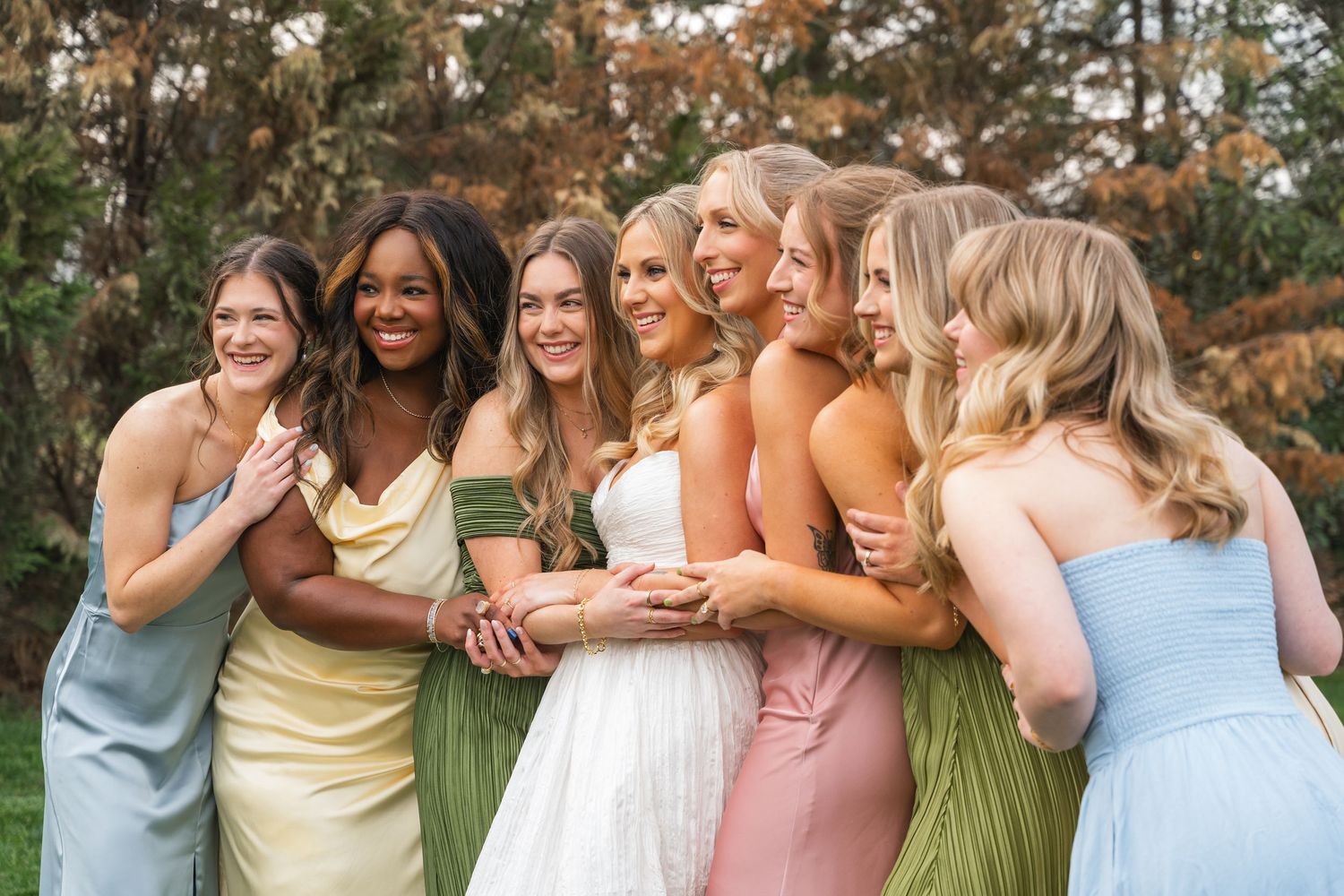 Bride in white gown surrounded by smiling bridesmaids in colorful mismatched dresses outdoors.