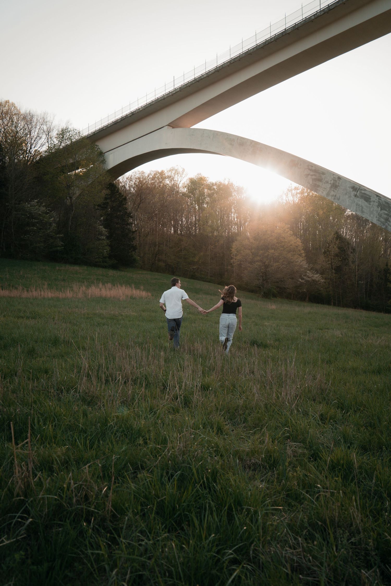 Couple holding hands walking through green field under arch bridge at golden sunset.