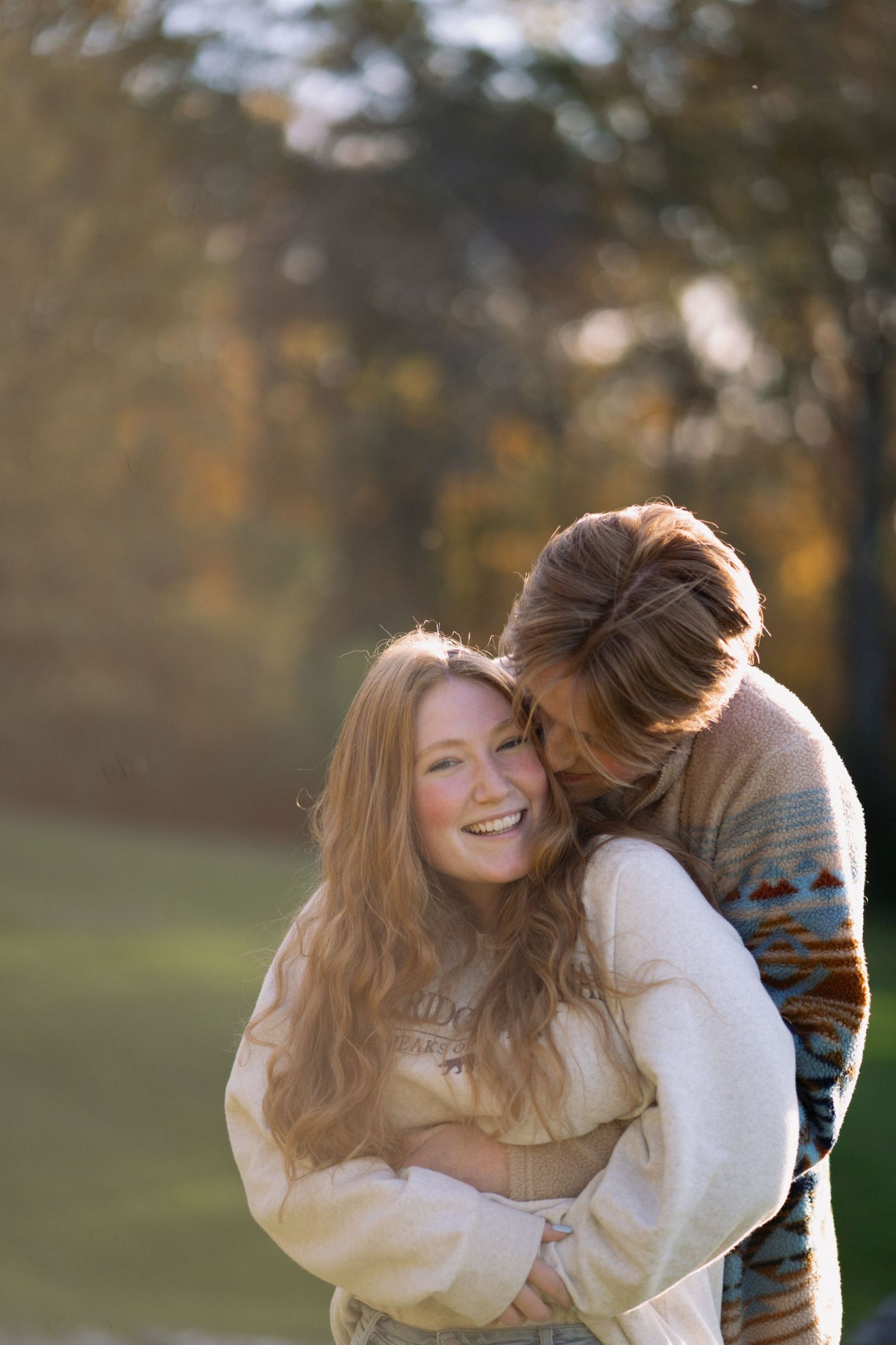 Couple embracing outdoors in autumn, woman with long red hair laughing joyfully while man hugs her from behind in warm sweaters.