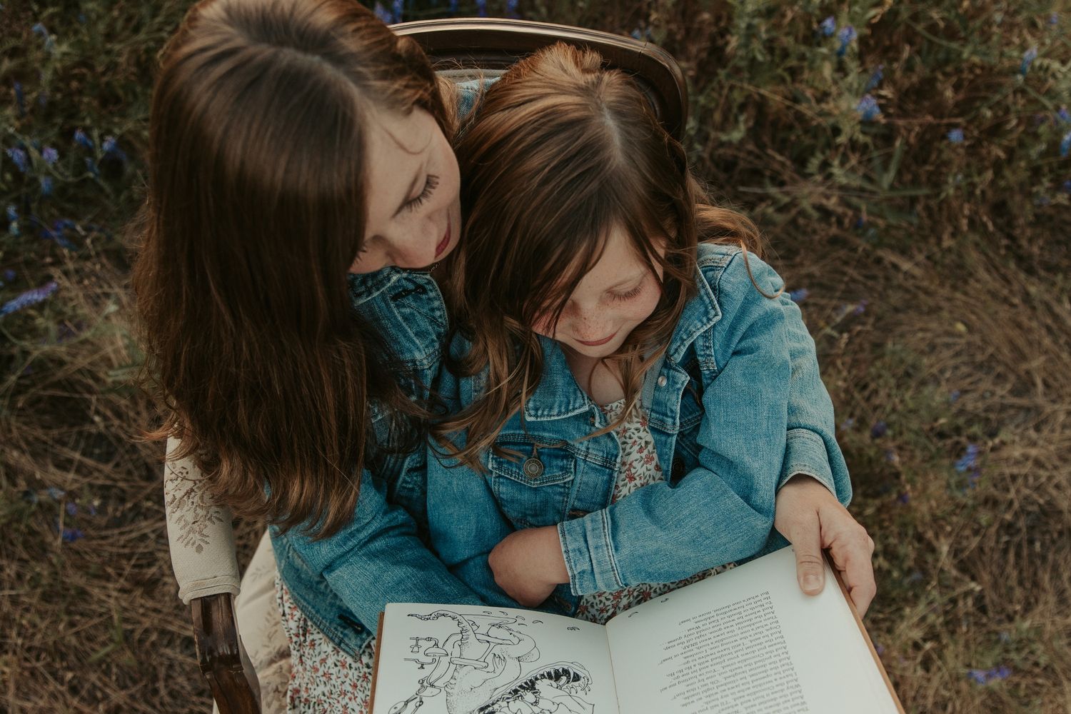 mom daughter dressed in matching jean jacket and floral dress sitting in purple flower field at sunset reading book with