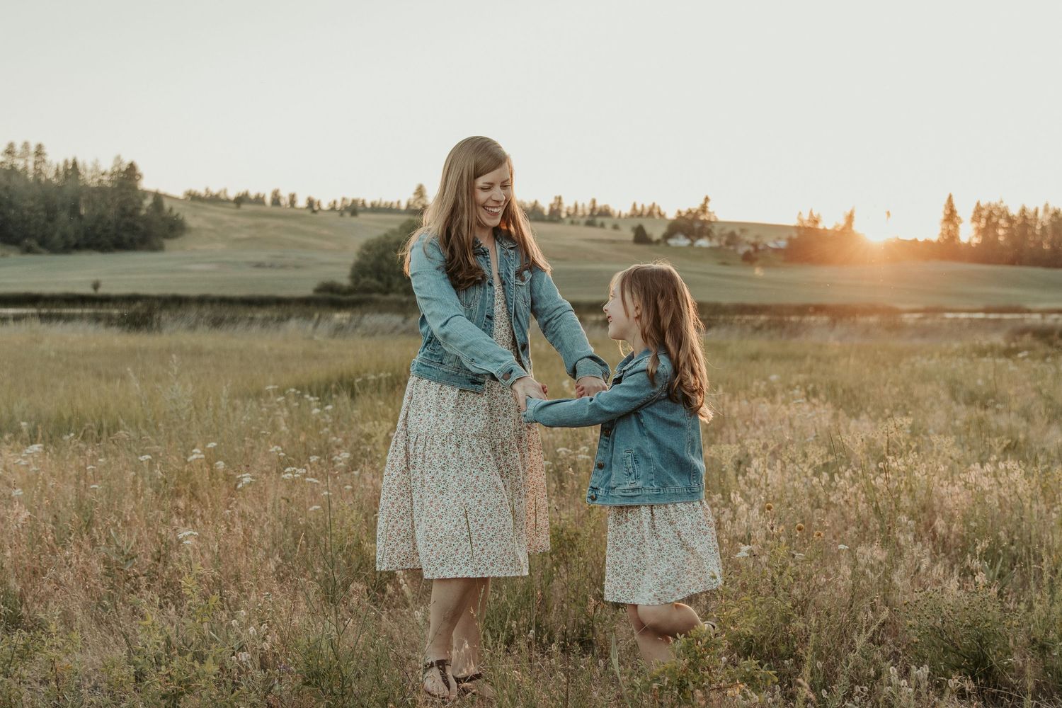 mom daughter dressed in matching jean jacket and floral dress dancing in a flower field at sunset with spokane photograp