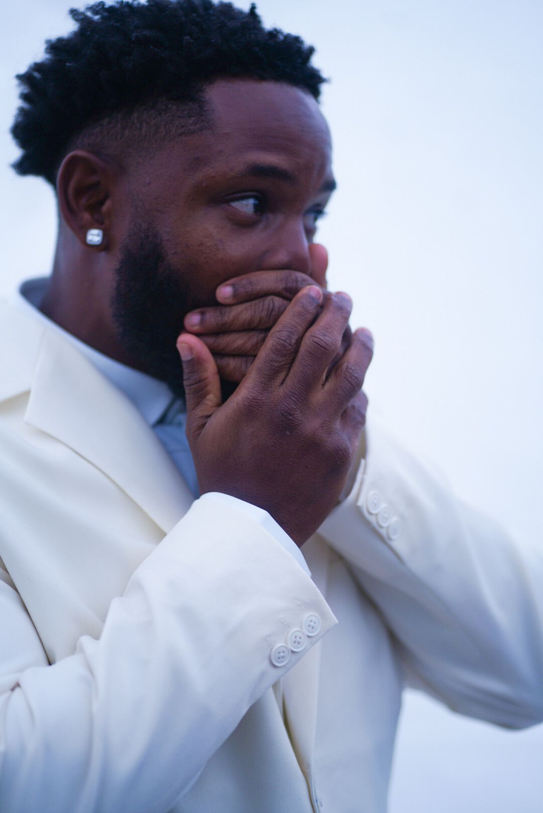 Emotional groom in white suit covering his face during wedding ceremony.