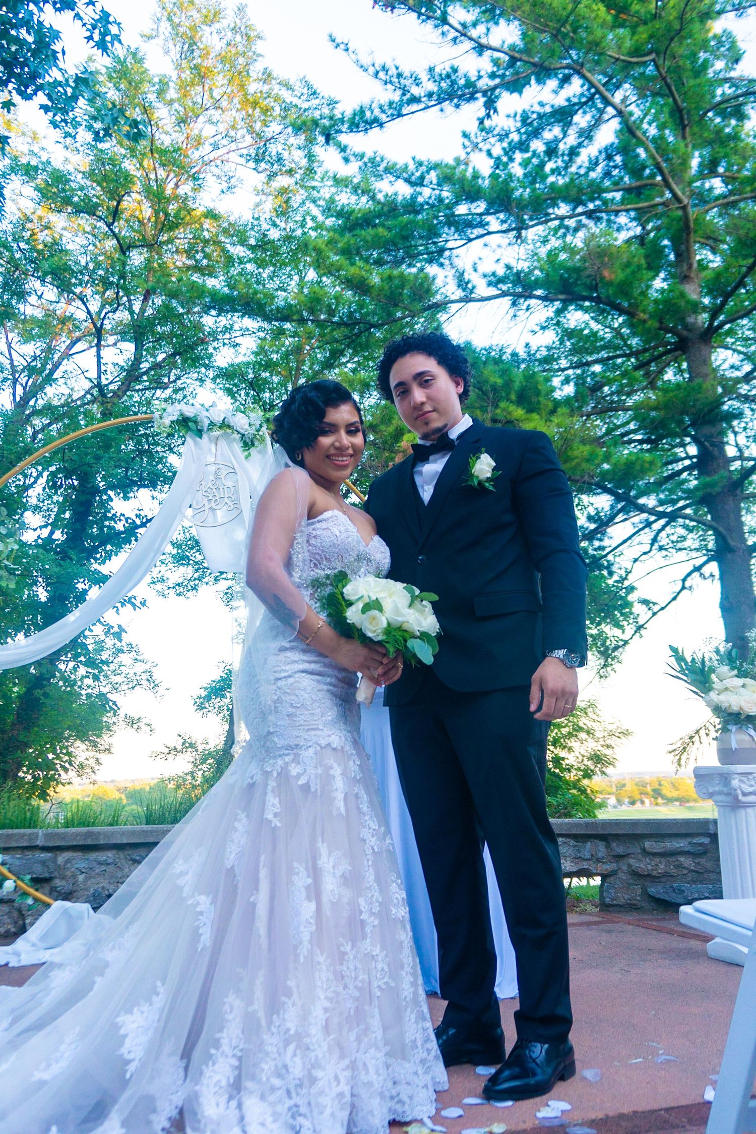 A newlywed couple poses for romantic photos under trees with white decorative elements.