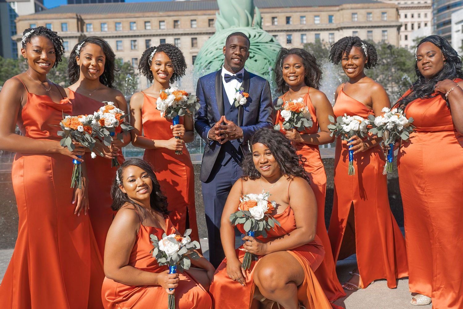 Wedding party in orange bridesmaid dresses and navy suit pose together outdoors with floral bouquets.