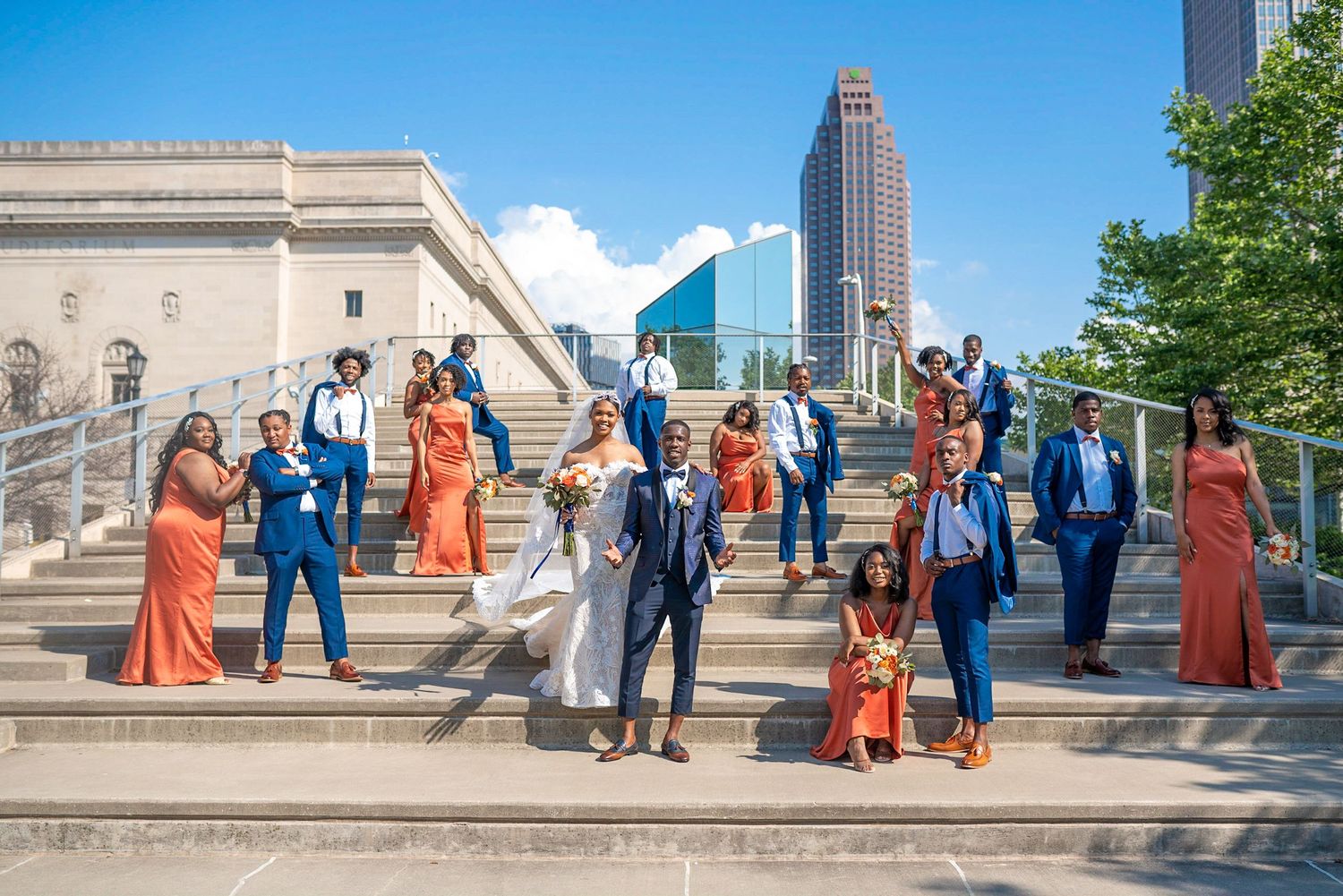 Wedding party members in orange dresses and blue suits pose on urban steps with skyscraper view.